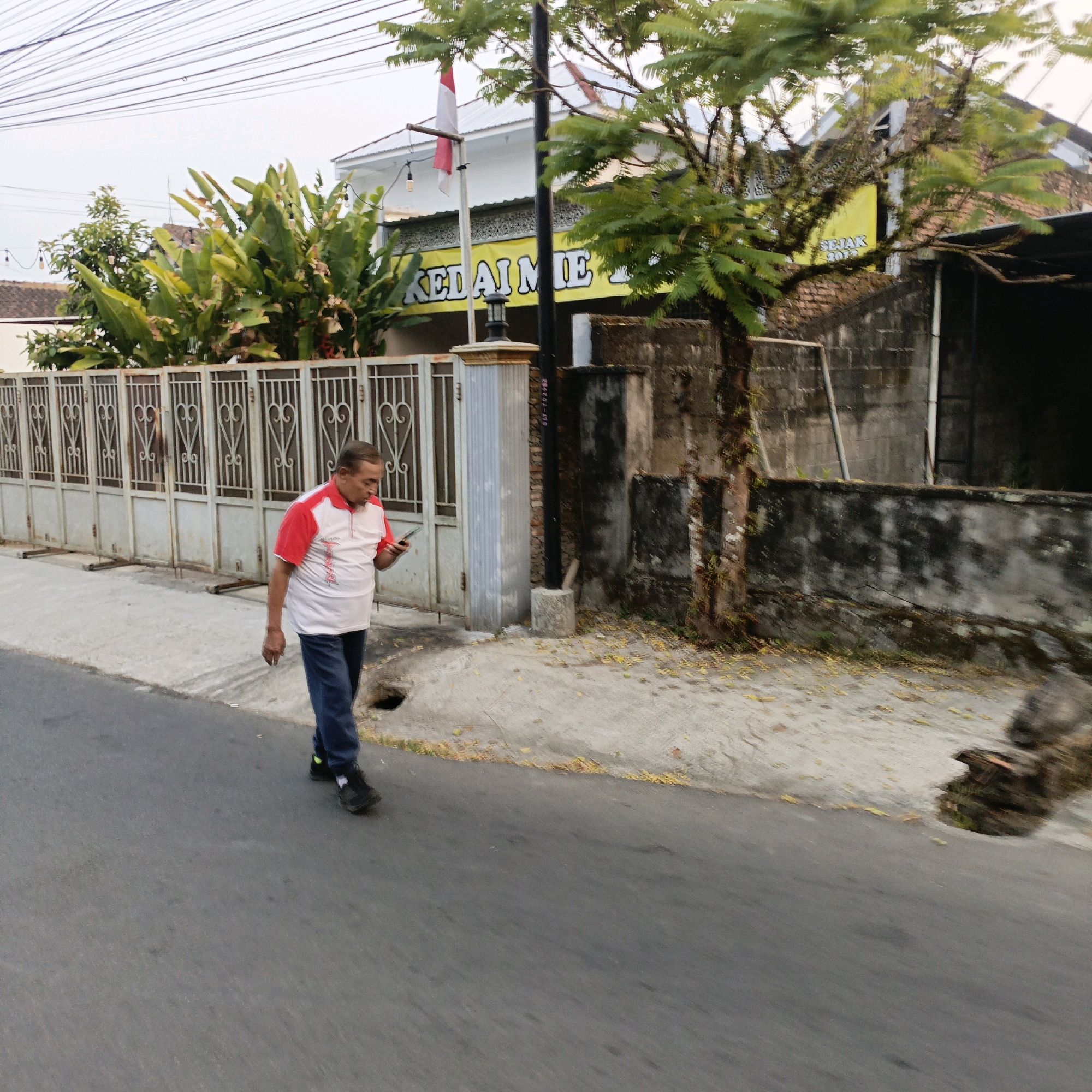 A middle-aged man with short, dark hair and a light complexion is walking on a paved road in front of a metal fence and a building. He is wearing a white polo shirt with red accents on the collar and sleeves, and dark blue pants. He is looking down at his smartphone, which he holds in his right hand. The man is positioned in the middle ground of the image, and the road extends into the foreground. Behind him, a tall metal fence with decorative patterns encloses a property with lush green plants and a building with a white facade and a red-tiled roof. A yellow banner with text is visible on the building. The sky is overcast, and there are power lines stretching across the top of the frame. The scene suggests a residential or commercial area in an Indonesian city, possibly Salatiga given the provided context.