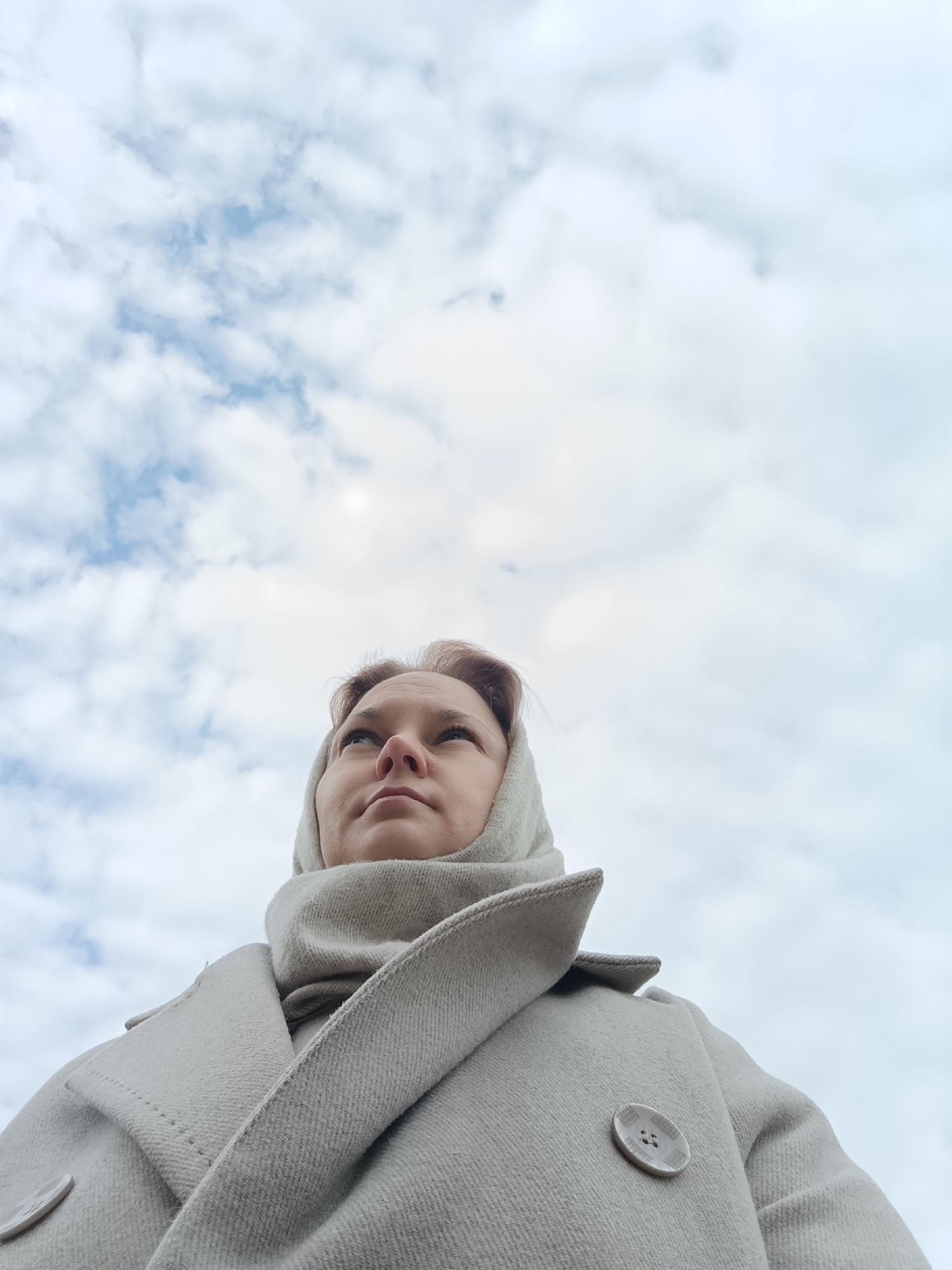 A low-angle shot depicts a woman against a cloudy sky. She has short, light-colored hair and is wearing a light grey coat. The coat is buttoned with a round, patterned button visible on the right side. She has a light grey scarf or head covering draped around her head and neck. The woman is looking upwards. The sky is filled with white and grey clouds.
The location is most likely not Марковский, Russia as the image contains no geographical information to confirm this.