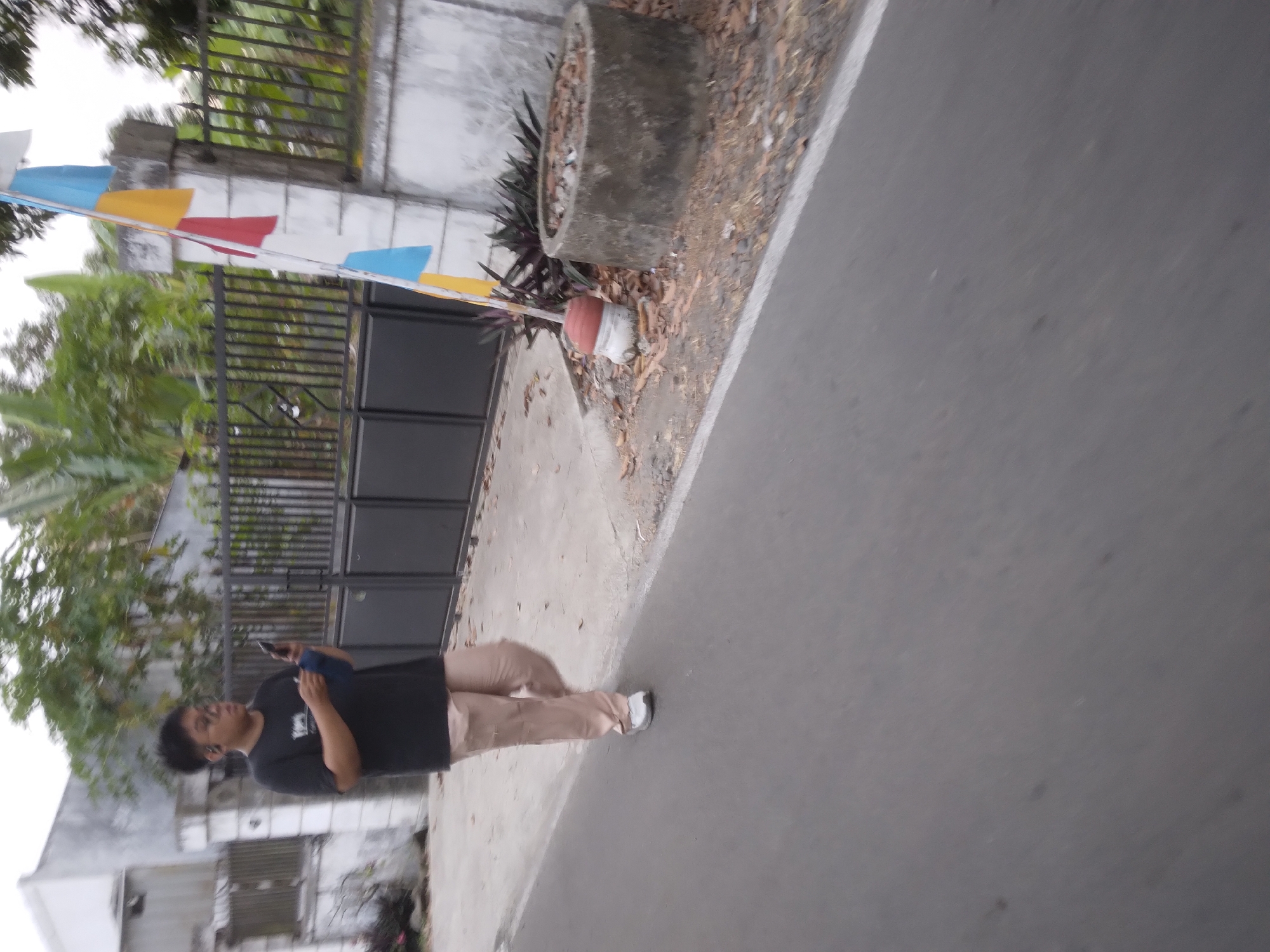 A young Indonesian male with dark, short hair and glasses walks down a paved road in Desa Pamijen, Kecamatan Sokaraja, Indonesia. He wears a black t-shirt and light brown pants, and is holding a dark-colored mobile phone and a blue cloth in his hands. He appears to be wearing earbuds. The road is bordered by a sidewalk with fallen leaves and some vegetation. Behind the sidewalk is a black metal fence, beyond which is a building with a white facade. A flag with horizontal stripes of white, yellow, red, and blue is attached to a pillar near the fence. To the right of the pillar, a circular concrete structure, possibly a well or planter, sits on the ground. The overall scene suggests an outdoor environment in a residential or semi-rural area.