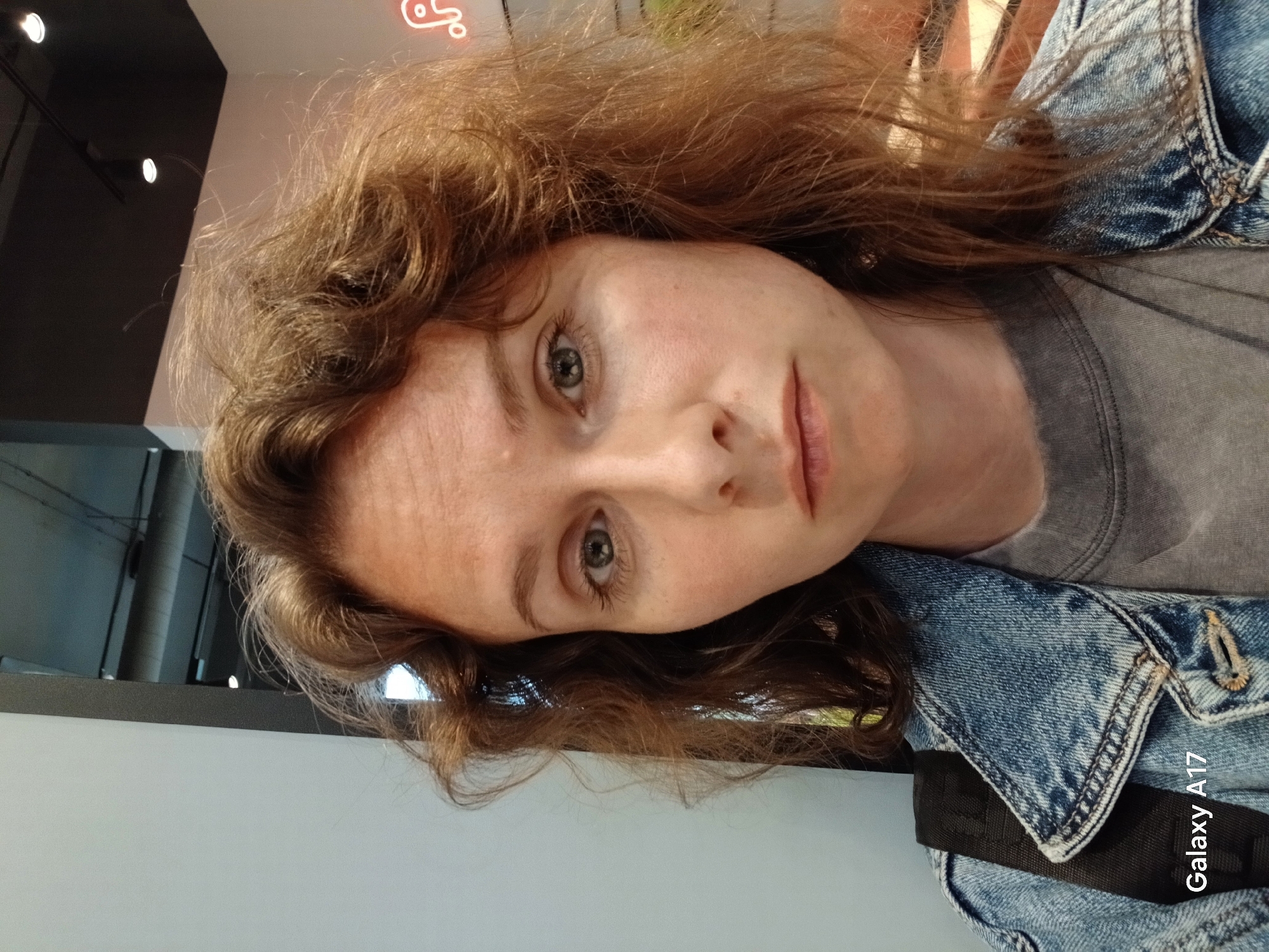 A front-facing portrait captures an individual with fair skin, light-colored eyes, and reddish-brown, wavy hair, looking directly at the camera with a neutral expression. They are wearing a blue denim jacket with a frayed collar over a plain grey crew-neck t-shirt. A black backpack strap is visible over their left shoulder. The background indicates an indoor commercial or public establishment, featuring a modern industrial aesthetic. To the viewer's left, a plain white wall is visible. The upper background shows a dark ceiling with exposed grey pipes/ducts and two visible track lighting fixtures. On the right side of the background, a partial red neon sign, resembling the curved part of a letter like 'P', is visible. The bottom left corner of the image features a "Galaxy A17" watermark. The scene is situated within the city of Yekaterinburg, Russia.