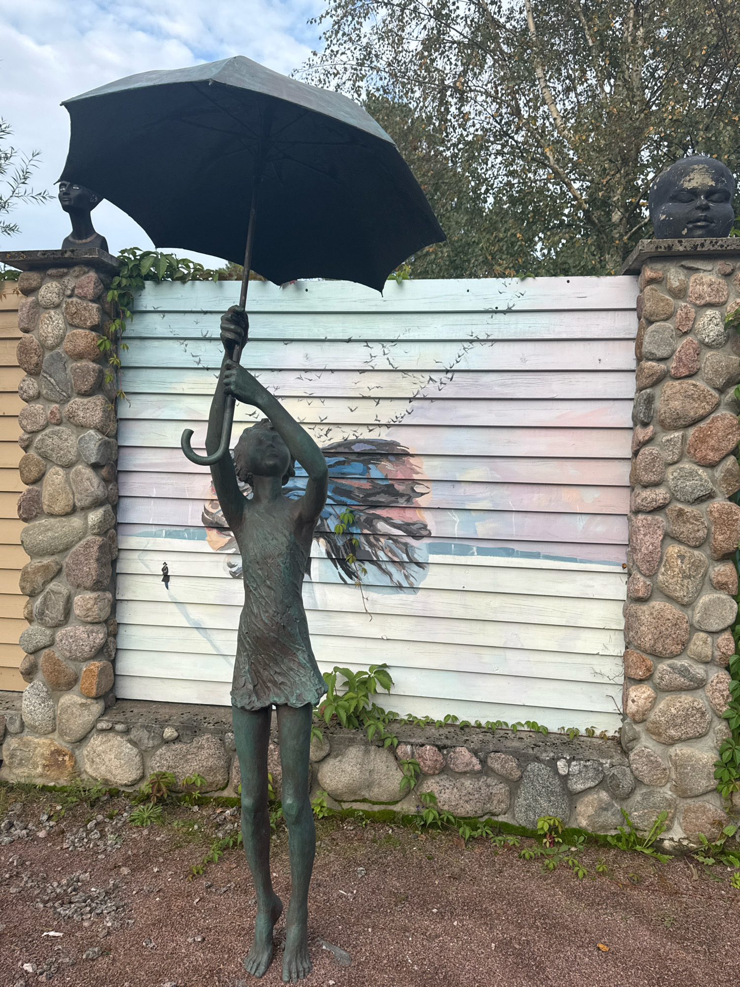 A bronze statue of a young girl holding an open umbrella stands in front of a wooden fence with a painted mural. The statue depicts the girl with her head tilted upwards, her arms raised holding the umbrella, and her feet bare. The mural on the fence features a hazy depiction of a face with flowing hair, surrounded by a flock of birds in flight and a pastel sky with hints of pink and blue. The fence is flanked by stone pillars, one of which is topped with a dark-colored, stylized head sculpture. Green vines grow along the fence and pillars. The ground in front of the fence is covered with reddish-brown gravel. The background shows trees and a cloudy sky. The location is identified as Интурист Снт, Russia.