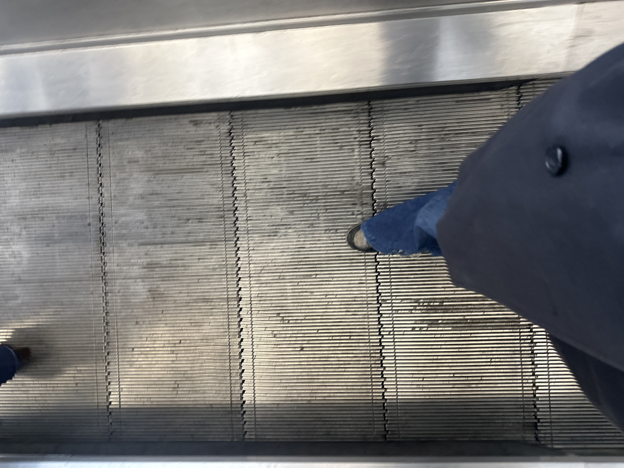The image provides a high-angle, overhead view of a person standing on a metal escalator or moving walkway, consistent with an indoor public environment such as a transit station, airport, or shopping mall in London, United Kingdom.
The primary subject is the lower half of a person. The individual is wearing blue denim jeans and a dark blue or black coat or jacket, identifiable by a single dark button visible on its fabric near the right leg. Dark-colored shoes are visible on both feet, with the left foot partially obscured and positioned further back on the moving surface.
The main object is the escalator or moving walkway, distinguished by its ribbed metallic surface composed of numerous parallel slats. These slats show signs of wear and some accumulated grime, suggesting frequent use. The surface exhibits a slight motion blur, particularly on the left side, indicating the movement of the walkway. The upper and lower edges of the frame show the smooth, polished metal sides or handrail structures of the escalator.
The scene is brightly lit, suggesting an indoor daytime setting, possibly with overhead artificial lighting or reflective surfaces bouncing ambient light. The metallic elements reflect light, creating a somewhat shiny appearance. The activity captured is the person being transported passively by the moving walkway. No text is visible anywhere in the image. The overall impression is one of mundane urban or travel transit.
