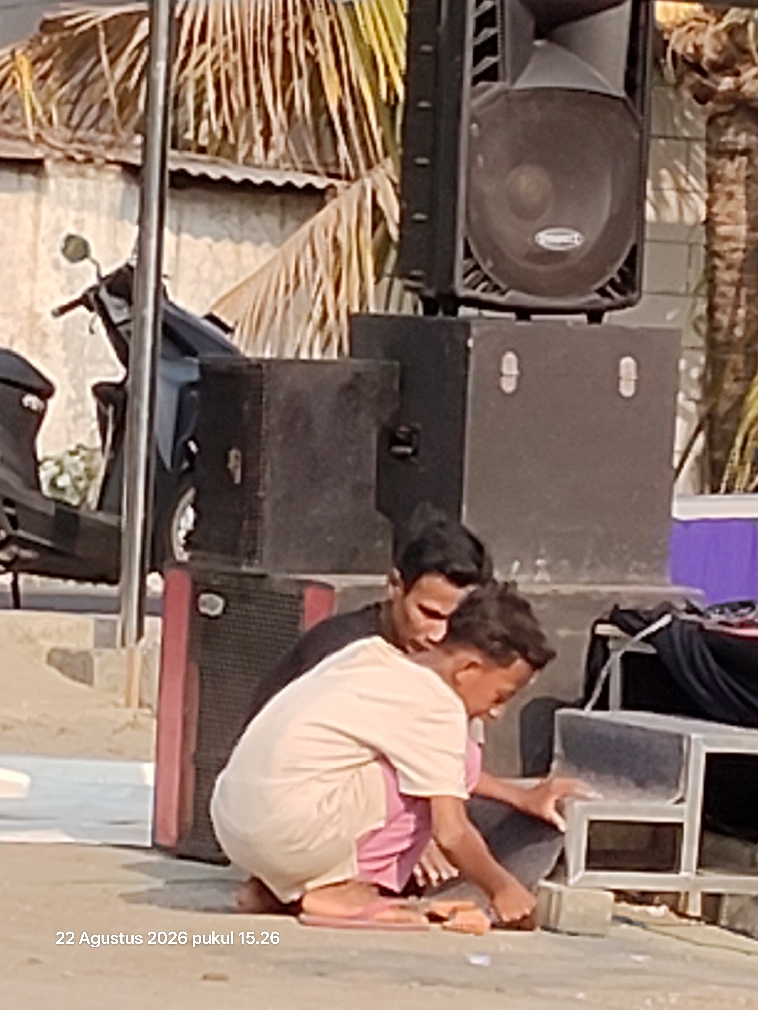 The image depicts two young males sitting on the ground in front of large speaker cabinets. The individual on the left is wearing a light-colored t-shirt and pink shorts, while the individual on the right is wearing a dark t-shirt and appears to be interacting with the speaker setup. The environment suggests an outdoor setting, possibly a street or public space, with palm fronds visible in the background and a motorcycle parked to the left. The overall scene appears casual, with the individuals engaged in some activity related to the sound equipment. The date and time "22 Agustus 2026 pukul 15.26" are overlaid on the bottom left of the image, indicating it was taken in Wedi, Indonesia.