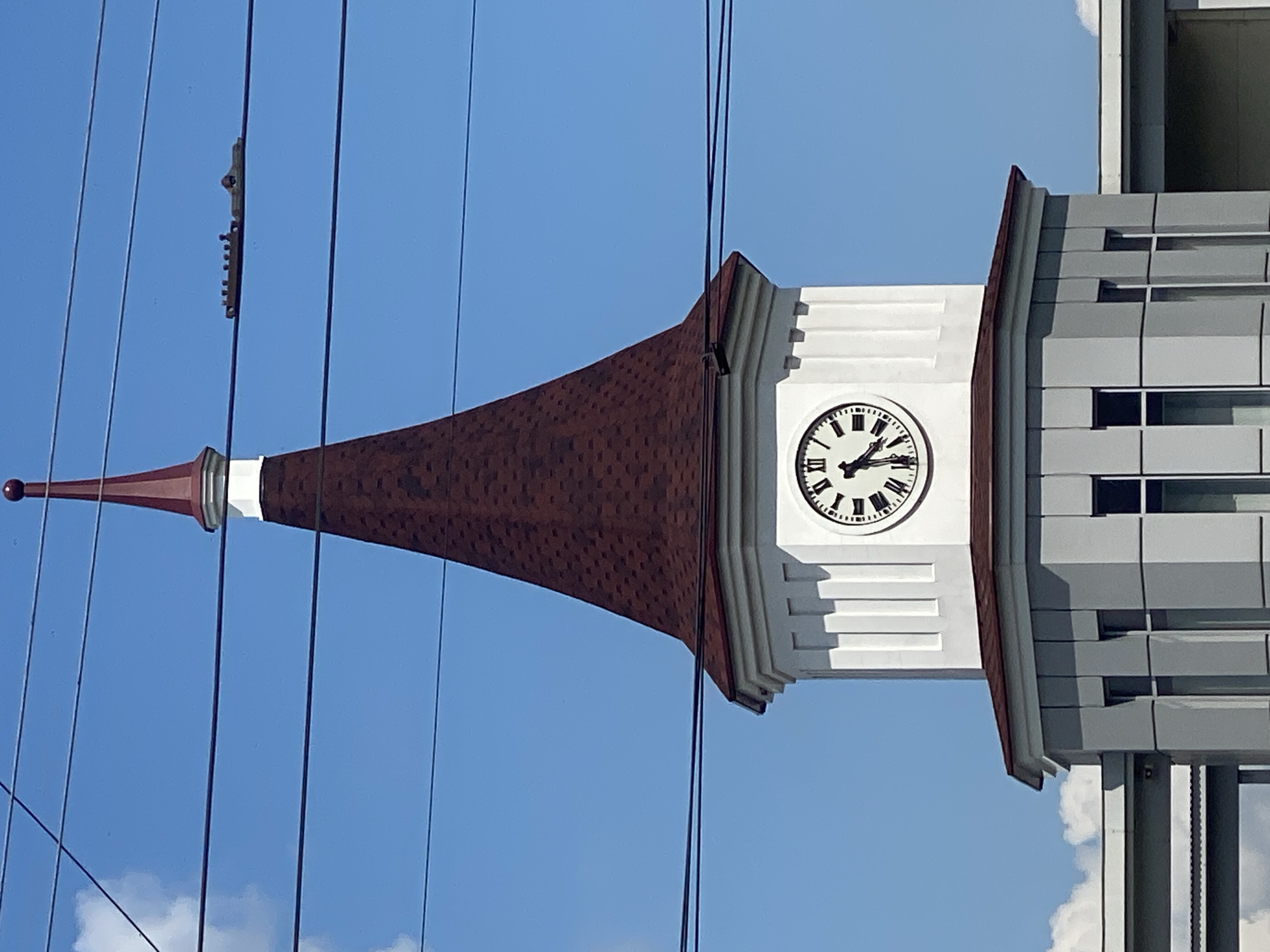 The image shows a close-up of a clock tower in Novokuznetsk, Russia, against a bright blue sky with scattered clouds. The tower has a white octagonal base with decorative vertical indentations, topped by a red, conical roof. A round clock face with Roman numerals is visible on one side of the tower, showing the time as approximately 10:47. Multiple dark power lines stretch across the frame in the foreground. The lower portion of the image reveals a grey building with vertical windows, suggesting this is part of a larger structure. There are no people visible in the image.