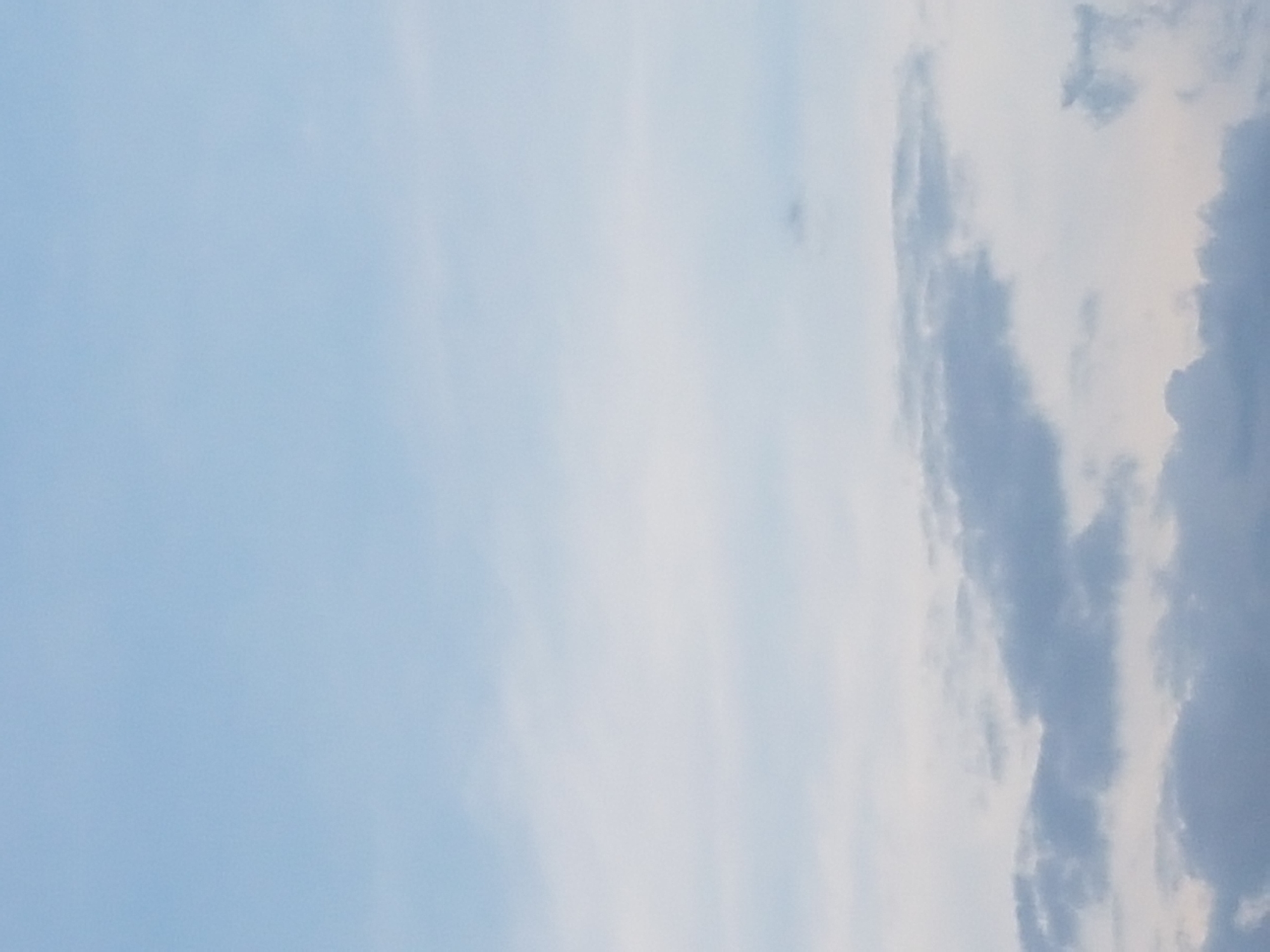 The image shows a sky with scattered clouds. The clouds are varying in shade from white to gray, with some appearing darker and more defined than others. The sky itself is a pale blue, lighter at the top and becoming slightly grayer towards the horizon. There are no discernible landmarks or structures visible, suggesting an open sky view. The image appears to be taken from an outdoor location.
