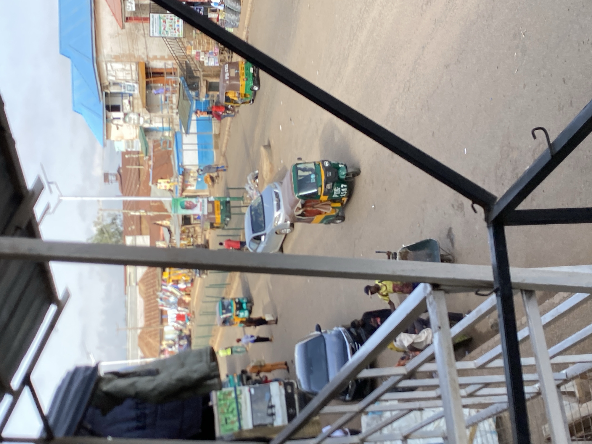 From an elevated perspective, a bustling street scene in Bukuru, Nigeria unfolds. A variety of vehicles, including a distinctively decorated tuk-tuk, traverse the asphalt road. Pedestrians are scattered along the roadside, suggesting a marketplace or commercial area. Buildings with diverse architectural styles line the street, some bearing colorful signage. In the foreground, a metallic structure, possibly part of a building or balcony, partially frames the scene, adding a layer of depth to the composition.