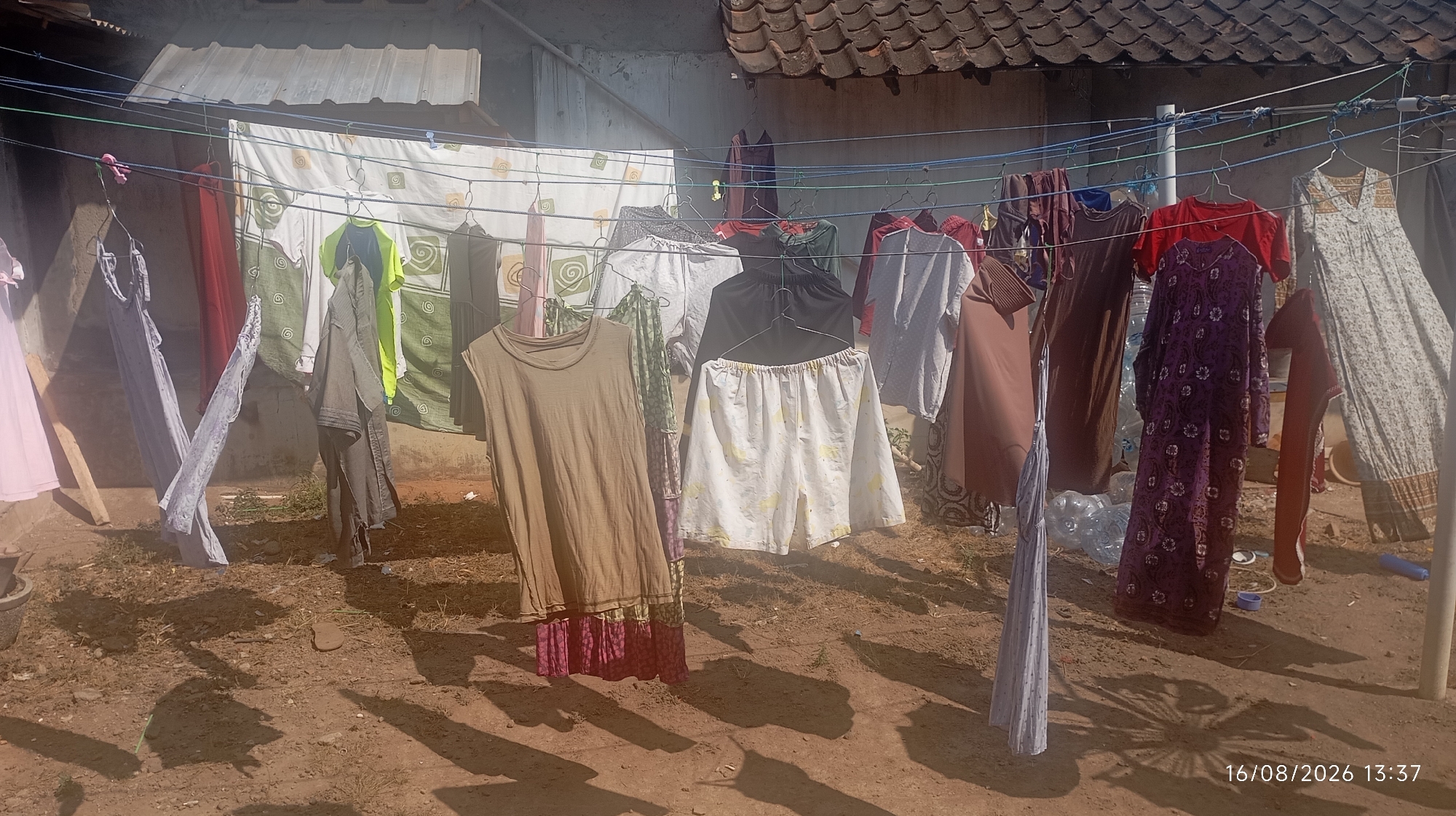 The image depicts an outdoor clothesline with various garments hanging to dry in what appears to be a residential area in Jatisobo, Indonesia. The setting is rustic, with a dirt ground and a tiled roof structure in the background. The clothesline is strung with multiple lines and is populated with a diverse assortment of clothing, including dresses, shirts, skirts, and possibly pants, in various colors and patterns. Some items are on hangers, while others are directly clipped to the lines. The sunlight casts distinct shadows on the ground, suggesting it is daytime. There are no people visible in the frame. The overall impression is one of everyday domestic life in a rural or semi-rural Indonesian environment.