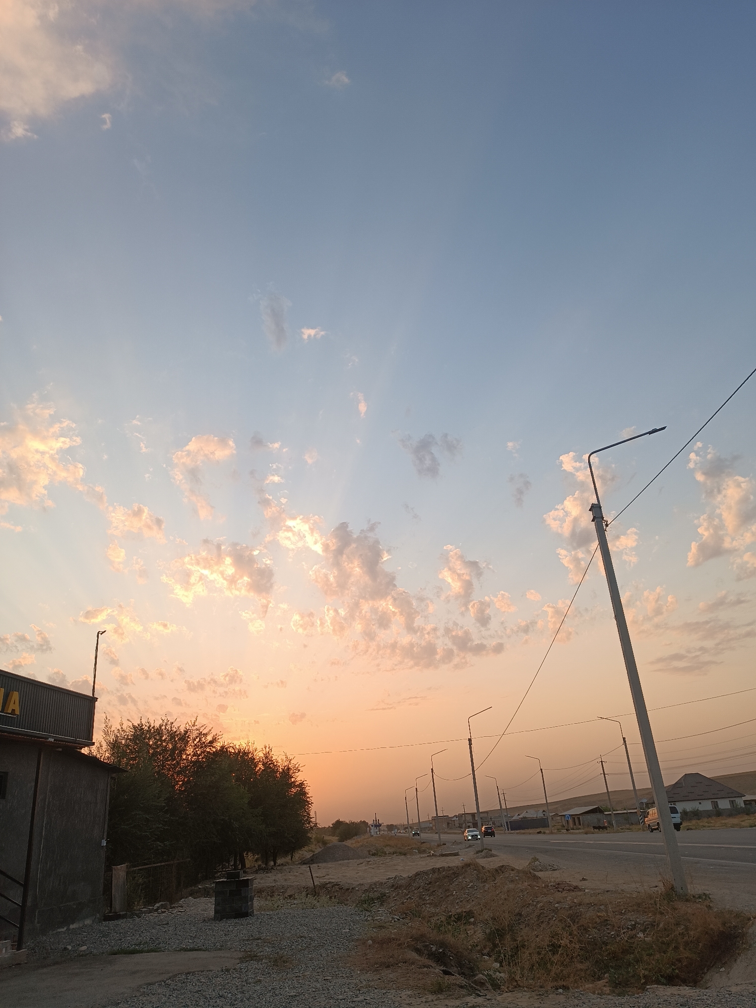 The image depicts a roadside scene in Jangy-Akman, Kyrgyzstan during sunset. The sky is a gradient of pale blue and soft orange, with scattered clouds tinged with pink and yellow. Several streetlights line a paved road that curves into the distance. The road is flanked by dry, sparse vegetation and some construction materials. On the left side of the image, a small building with a dark facade and a yellow sign is visible. Trees with dense green foliage stand behind the building. A few cars are driving on the road, and in the background, houses with slanted roofs can be seen against the arid landscape. A single utility pole with wires stretches across the sky. The overall atmosphere is calm and rural, characteristic of a sunset in a developing area.