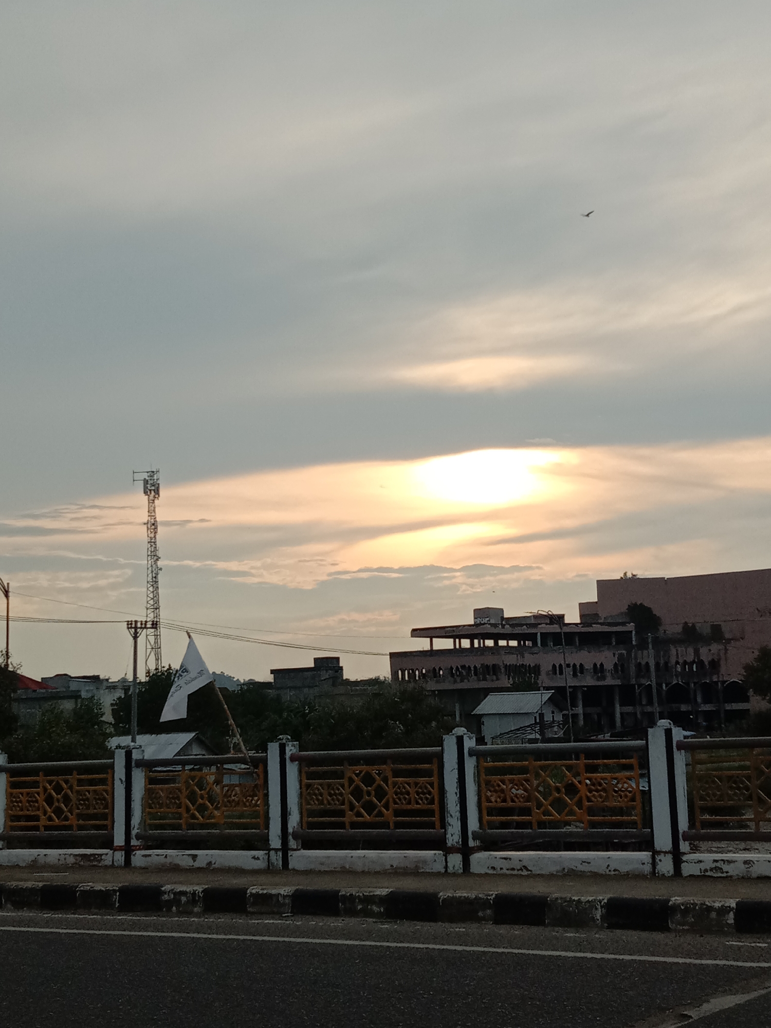 The photograph captures a cityscape in Lhokseumawe, Indonesia, under a sky with sunlit clouds. The view is taken from what appears to be an elevated position, likely a bridge, judging by the road and bridge railing with a patterned yellow design visible at the bottom.
In the midground, a mix of structures including a tall cell tower, smaller buildings, and what seems to be a larger, multi-story, partially constructed or derelict building can be seen. A flag is waving. Vegetation is visible amongst the buildings.
In the sky, there's a single bird flying. The light indicates either sunrise or sunset, casting a glow on the underside of the clouds.
