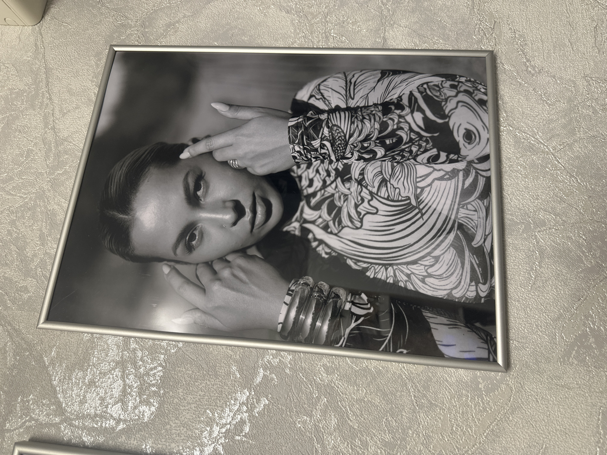 A framed black and white photograph of a woman is displayed on a textured wall. The woman is positioned in the center of the frame, looking directly at the viewer with her fingers placed on her temples. She is wearing a long-sleeved patterned top and multiple bracelets on her left wrist. The photograph is framed in a silver-colored metal frame. The wall behind the photograph has a raised, stucco-like texture.