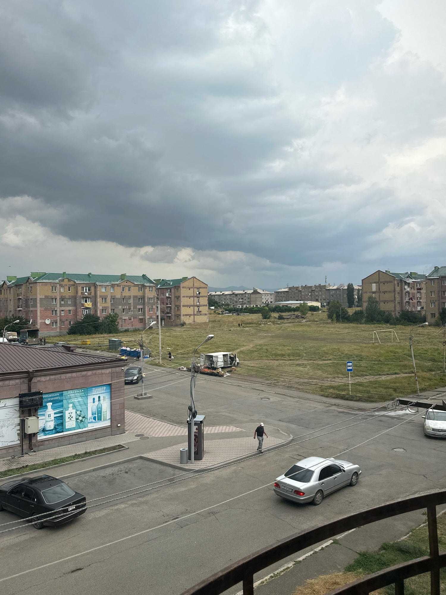 This is an exterior, eye-level, slightly angled, full shot of a street scene in Gyumri, Armenia, on a cloudy day. The foreground features a railing, suggesting the shot was taken from a balcony. In the mid-ground, there is a street with several cars, including a black sedan, a silver sedan, and a silver car parked on the right. A pedestrian, wearing a white cap and dark clothing, walks along the sidewalk near a payphone and trash receptacle. To the left, a building with large advertisements is visible. Beyond the street and sidewalk, a large, open field is present, with a soccer goal frame in the distance. Several apartment buildings, characterized by their yellow and gray brick facades and green roofs, line the horizon, interspersed with trees and other structures. The sky is overcast with heavy, gray clouds.