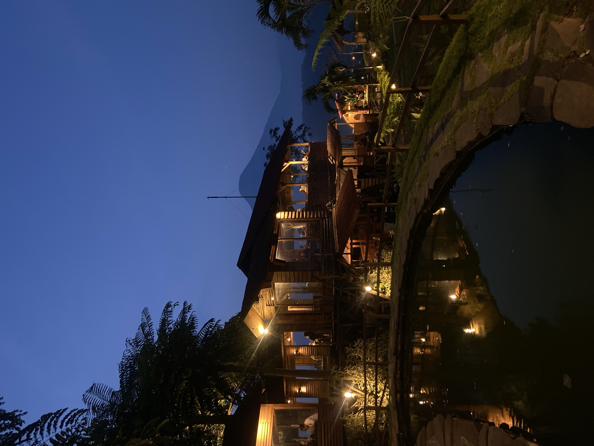 The image depicts a multi-story wooden structure, possibly a restaurant or lodging, illuminated at night. The architecture appears to be traditional Indonesian. The structure is reflected in a body of water, likely a pond or a small lake. The presence of trees and vegetation indicates a natural or landscaped environment. People can be seen inside the building on different levels. A fence lines a pathway next to the water. The sky is dark blue, suggesting twilight or night. The overall atmosphere is peaceful and serene. The city of Tamiajeng, Indonesia, is noted as the potential location.