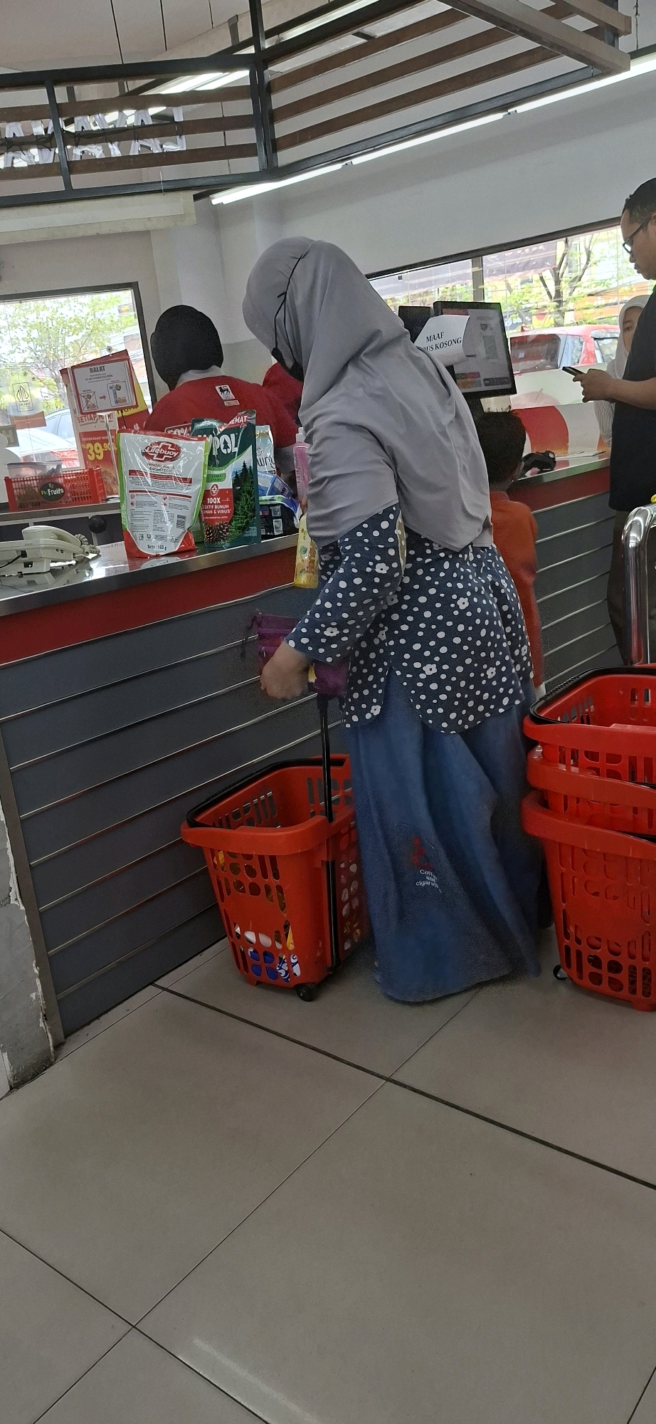 An outdoor scene, likely a convenience store, shows a woman in a light gray hijab and a navy blue polka dot shirt with a long blue skirt, using crutches. She is pushing a red shopping basket. She appears to be interacting with a cashier, whose red shirt is visible. Another person, wearing a dark shirt, is standing nearby looking at their phone. A child in an orange shirt is standing behind the woman. The scene is likely in Surabaya, Indonesia, given the context.