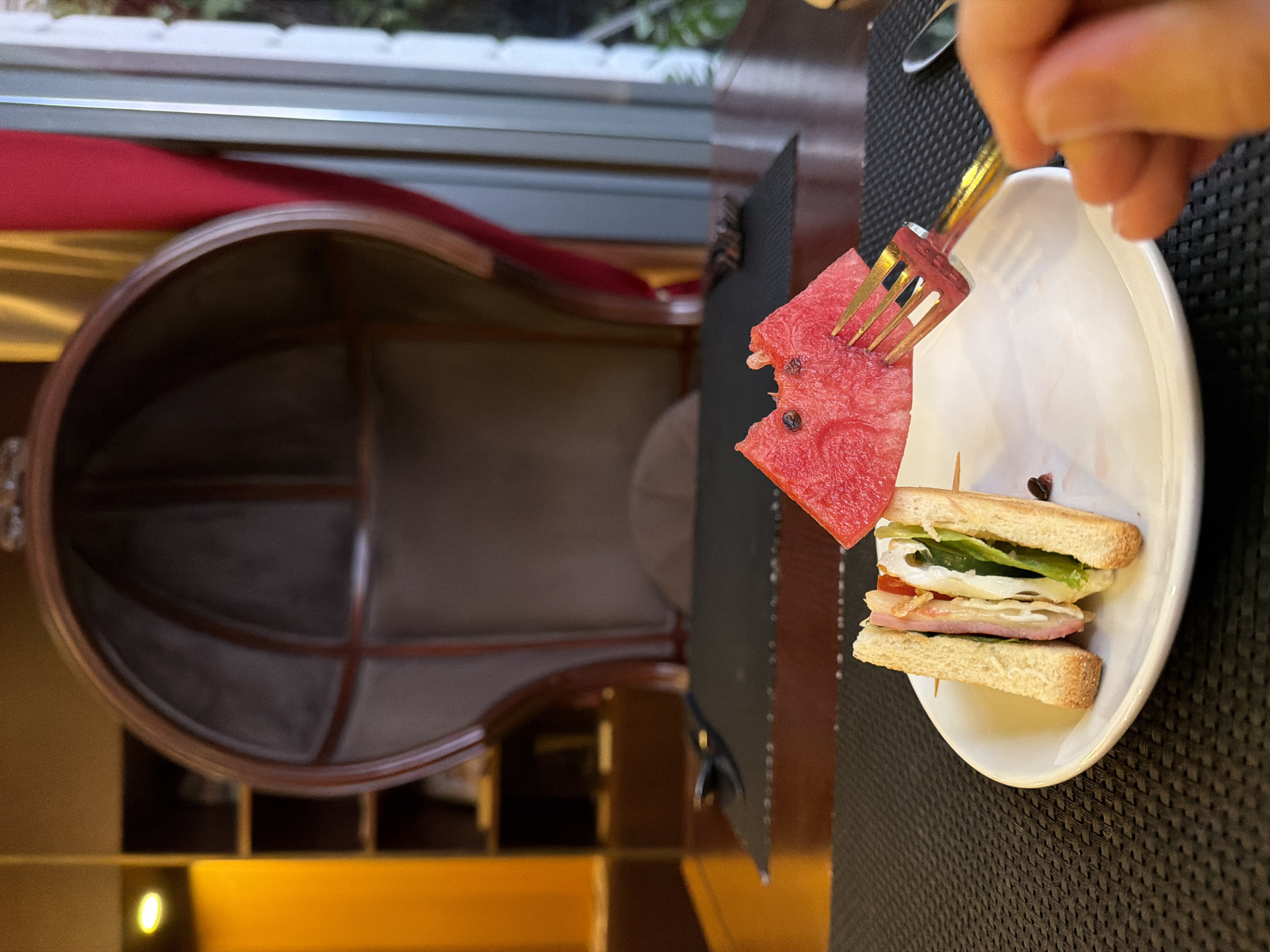 A close-up, eye-level shot shows a person's hand holding a golden fork as they reach for a slice of watermelon with a bite taken out of it. The watermelon slice is speared by a wooden skewer along with a small sandwich. The sandwich, cut in half diagonally, consists of toasted bread, lettuce, tomato, and a pink-colored meat, possibly ham or turkey. Both the watermelon and sandwich are on a white plate, which rests on a black placemat. The background features a large, ornate, brown chair with a high back, suggestive of a luxurious dining or lounge setting. Further in the background are shelves filled with items, a red curtain, and a window showing a glimpse of greenery. The overall impression is that of a meal being served in a well-appointed indoor space, possibly a restaurant or hotel lounge in Thuận Hóa Ward, Vietnam.