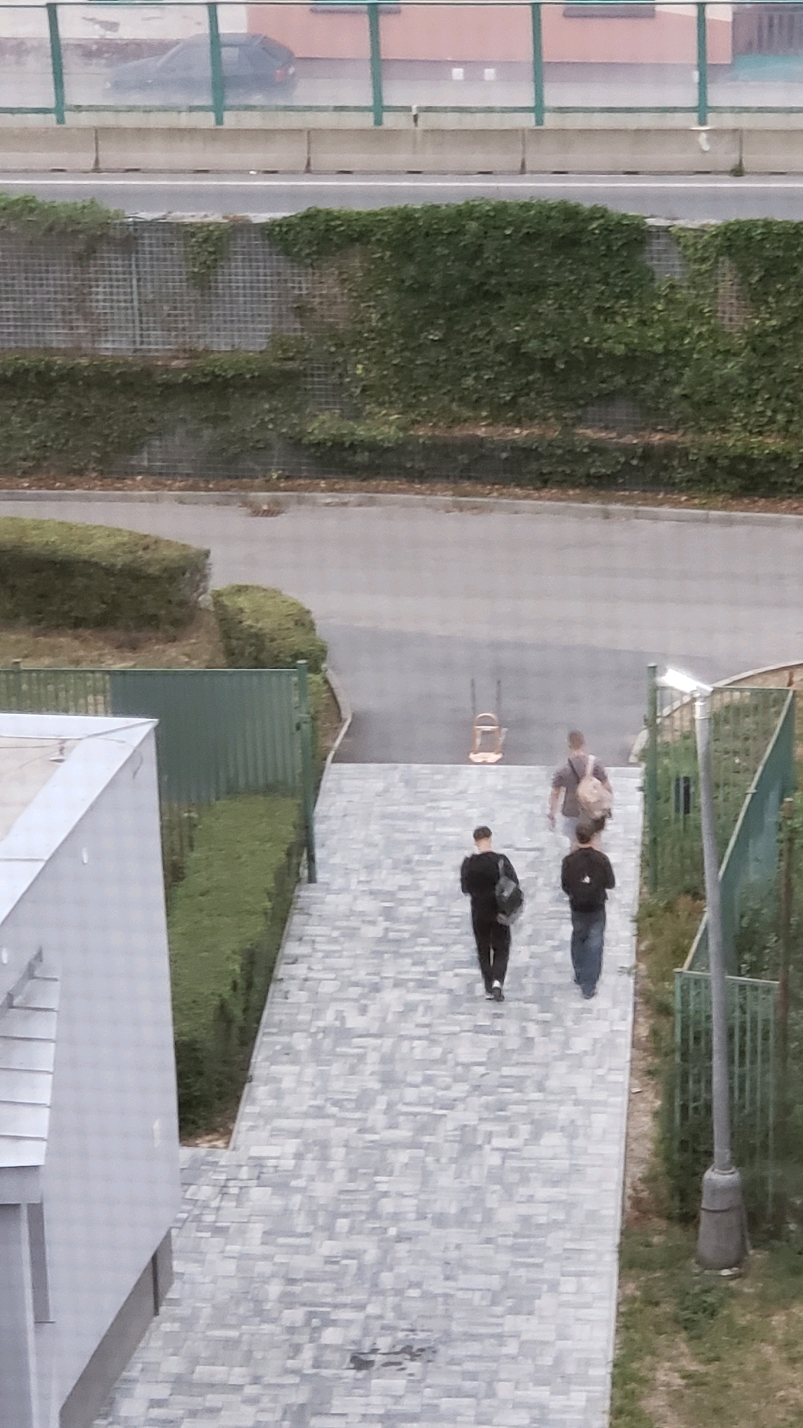 The image captures an outdoor scene, likely in the city of Prešov, Slovakia, viewed from an elevated perspective. In the foreground, a grey, paved walkway with a patterned brick design descends from the upper right to the lower left. Three individuals are walking down this path. The person in the front left is wearing a black outfit and carrying a backpack. To their right and slightly behind is another individual in a dark top and blue jeans, also with a backpack. Further up the path and to the right, a third person with light-colored hair and a backpack is walking away from the camera. To the left of the walkway, a modern building with a white facade and a grey roof is partially visible. Adjacent to the building and walkway is a manicured green hedge. Beyond the hedge, a green metal fence runs along a paved area with some scattered debris. In the background, a road with a visible lane marking is bordered by a retaining wall covered in dense green ivy. A dark-colored car is faintly visible on the road. The overall environment appears to be a public space with landscaping and architectural elements.