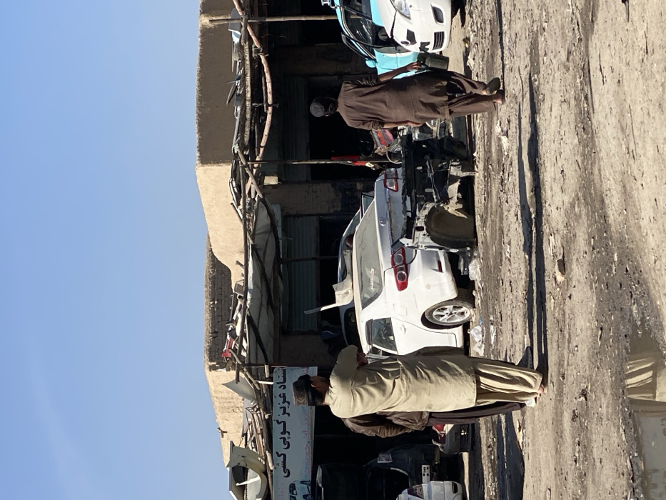 The scene depicts an outdoor auto repair and salvage yard in Kandahar, Afghanistan, specifically identified by a sign displaying "استاد عزيز كوبي كشي" (Ustad Aziz Auto Parts). The ground is unpaved, dusty, and strewn with debris, including a small puddle reflecting light in the foreground-left.
Three adult males are visible. In the center-left, one man stands facing right, wearing a light tan shalwar kameez and a dark cap, with his arms crossed. Partially obscured behind him is another man in a brown jacket. On the far right, a third man, dressed in a dark brown shalwar kameez and a dark cap, walks away from the viewer towards the background, carrying a dark object in his left hand.
The background features low-rise, earth-toned buildings with flat roofs piled with various discarded materials and scrap metal. Makeshift awnings extend from the building fronts.
Multiple vehicles are present. A prominent white sedan (likely a Toyota Corolla, ~2007-2013 model) occupies the center-midground, heavily damaged with its rear bumper and trunk area removed, exposing the chassis. A white shovel-like object rests on its roof. In front of this sedan, a disassembled dark vehicle frame sits on the ground. To the far right, a light blue/teal sedan with an open driver's door and a partially visible white sedan are parked, both appearing damaged or undergoing repair. The sky above is clear and light blue.