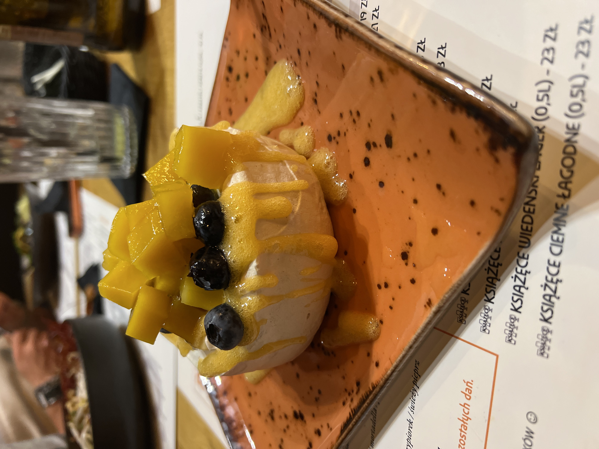 An overhead shot focuses on a dessert of meringue topped with diced mango and blueberries, drizzled with a mango sauce. The dessert rests on an orange, speckled, square plate. In the background, a glass of water and a bottle of beer are visible on a wooden table. A menu with Polish text is partially visible on the table. The overall setting appears to be a restaurant in the city of Białystok, Poland.