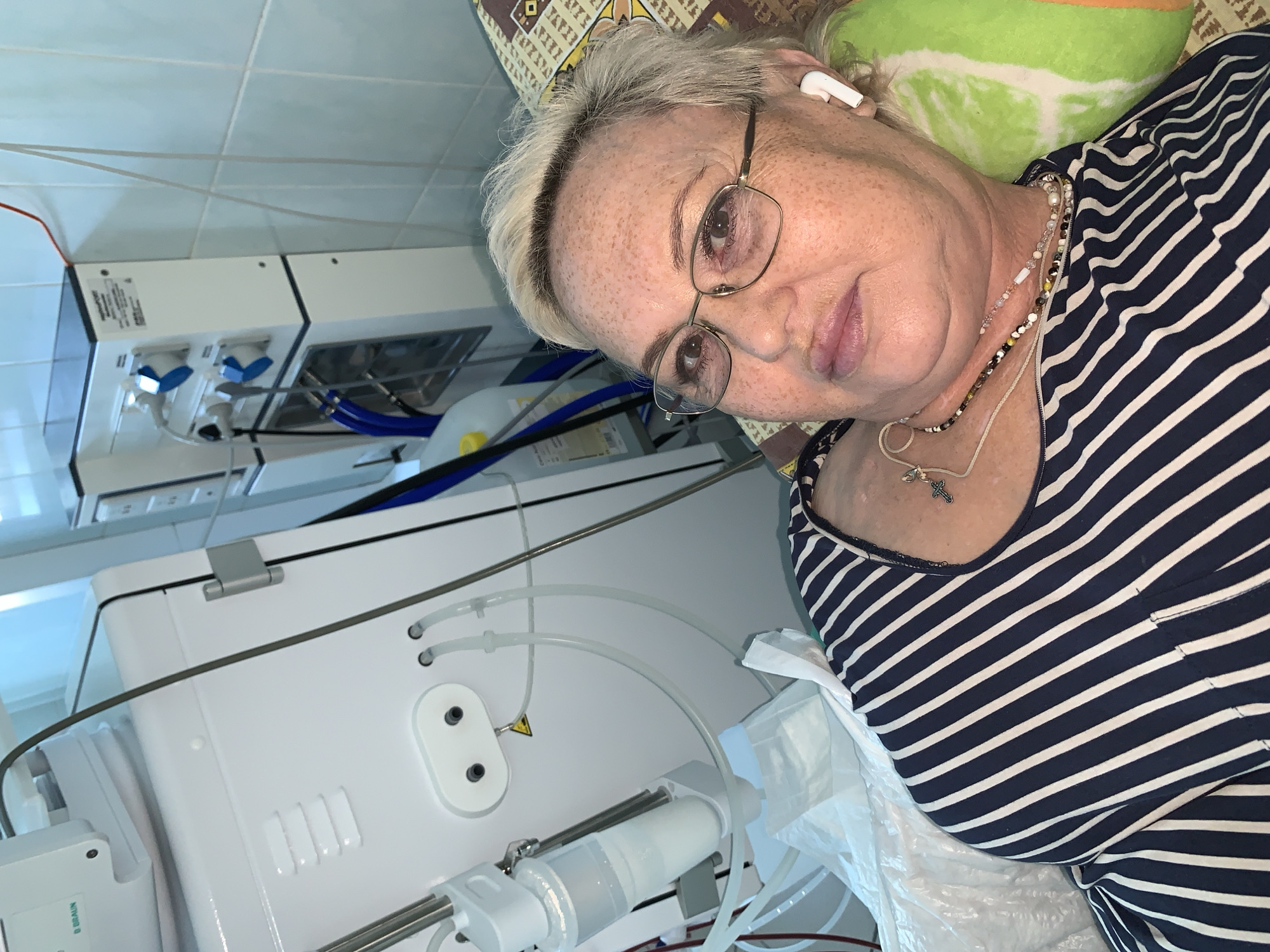The image shows a middle-aged Caucasian woman with blonde hair, wearing glasses, a striped shirt, and white earbuds, lying down in a medical setting. She appears to be undergoing a medical procedure, possibly dialysis, as indicated by the presence of medical equipment in the background, including a dialysis machine, tubes, and a IV bag. The room appears to be a hospital or clinic, with tiled walls and a bed. The woman is looking directly at the camera with a neutral expression. The location is specified as Chaikovsky, Russia.