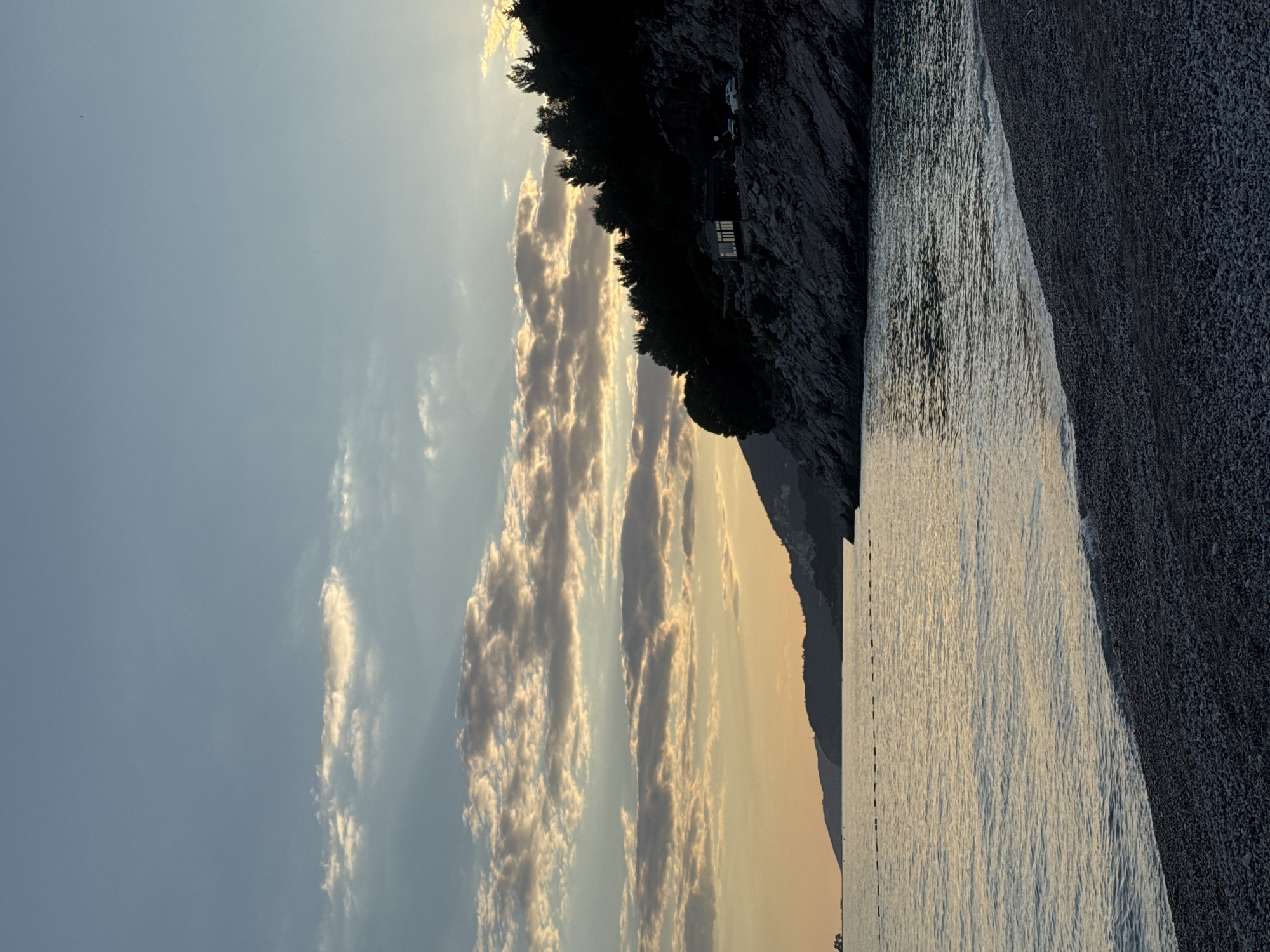 The image shows a view of Kamenovo beach in Montenegro, with the Adriatic Sea on the right and a rocky cliff covered in trees on the upper right. The sky is visible on the left, with clouds illuminated by the sunset. The beach consists of small pebbles. A structure appears to be built into the cliffside.