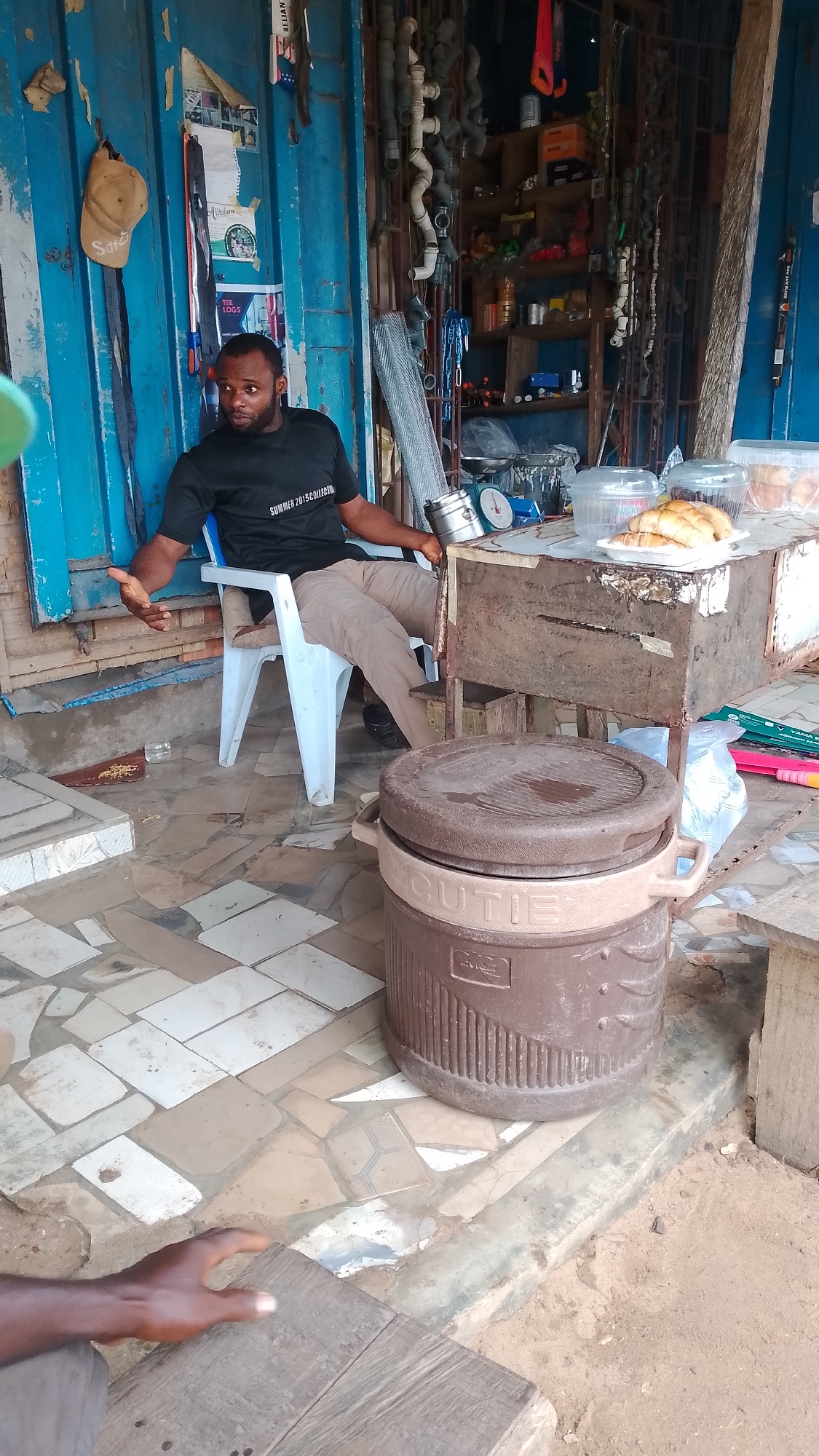 A man is sitting on a plastic chair in front of a shop in Asubi, Nigeria. The shop's facade is primarily blue, with shelves displaying various items, including tools, pipes, and hardware. The man is wearing a black T-shirt and khaki pants. He is holding a small, round object. There is a table next to him with food in containers. A large brown cooler with the word "CUTIE" on it sits in the foreground. The floor is a mix of tile and exposed earth. Another person's hand is visible in the lower left corner of the frame.