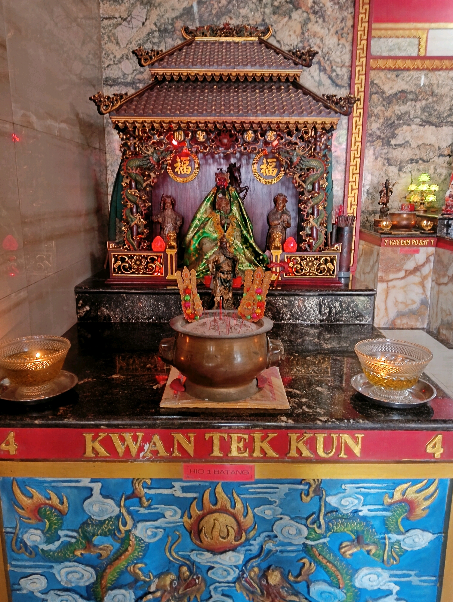 The image depicts an altar within a temple, likely a Buddhist or Taoist shrine, located in Rangkasbitung, Indonesia. The central focus is a tiered, ornate structure resembling a miniature temple, adorned with gold accents and intricate carvings of dragons and auspicious symbols like the character "福" (fú), meaning fortune. Within this structure, two statues of deities are visible, one draped in green fabric. In front of the altar, a brass censer filled with incense sticks and ashes sits on a wooden base. To the left and right of the censer, two glass bowls containing burning oil lamps are placed on saucers. The altar is situated on a dark, polished surface, possibly granite, and a red banner with gold lettering, reading "4 KWAN TEK KUN HIO 1 BATANG," is mounted above a vibrantly painted panel depicting dragons and clouds. The background consists of marble-patterned walls, and the overall lighting suggests an indoor setting, possibly with artificial illumination.