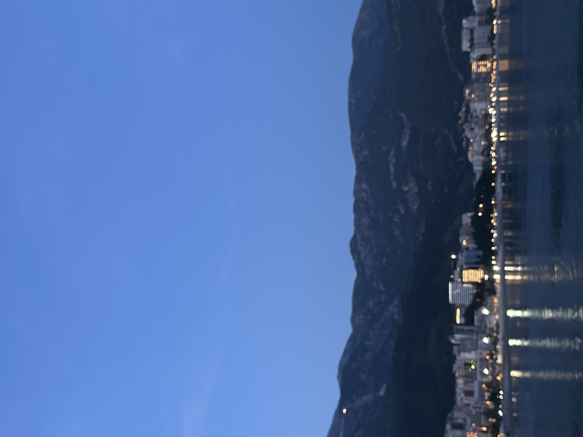 A nighttime shot in Boreti, Montenegro, shows a dark mountain range to the right, abutting a lighted cityscape. The buildings' lights reflect on the water in the foreground. The sky to the left is a gradient of blues.