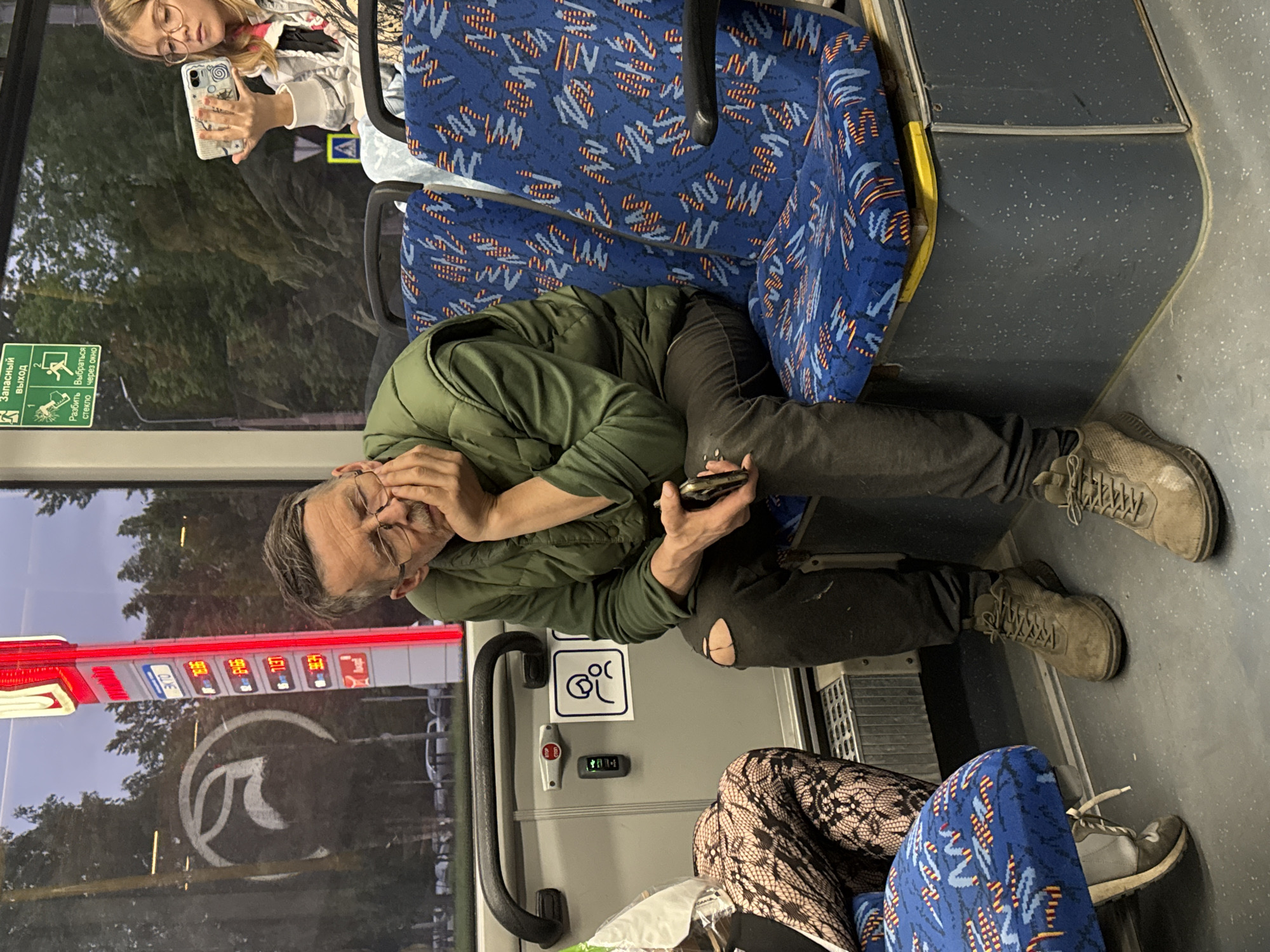 A man is seated on a bus in Saint Petersburg, Russia. He wears glasses, a green jacket, and dark pants with a hole in the knee. He holds a smartphone in his hands and his face is partially covered by his hand. Next to him, a person wearing fishnet stockings occupies the adjacent seat. The bus has blue patterned seats and a handicapped symbol is visible. Another passenger with blonde hair and glasses is visible towards the upper left corner, holding a smartphone. Through the bus window, a street is visible, with a sign displaying gasoline prices.