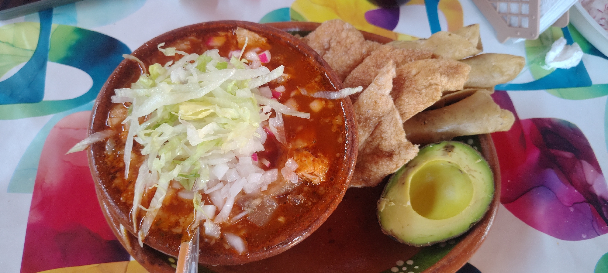 A top-down shot of a plate of Mexican food, likely pozole, in Ocotepec, Mexico. The main dish, served in a brown clay bowl, is a red broth with shredded lettuce, diced onions, and radishes. Alongside are fried tortillas, some puffed and crispy, others rolled and filled, plus half of an avocado. The plate rests on a colorful tablecloth with a pattern of abstract shapes and colors. A metal spoon is inside the bowl.