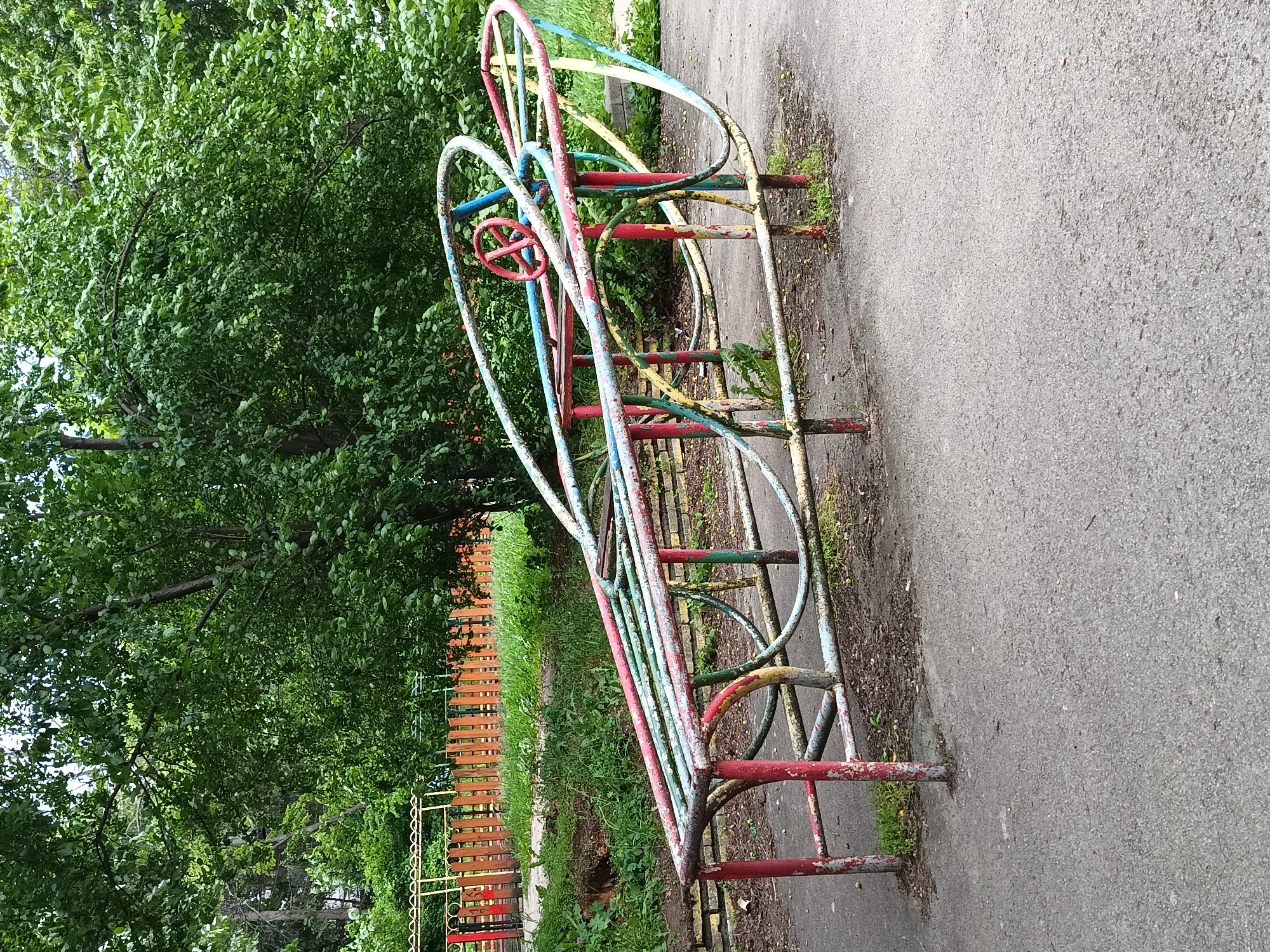 A children's playground is depicted in this outdoor shot. The scene features a weathered, metal climbing structure with multiple colored bars – red, blue, yellow, and green – set along an asphalt path. The playground structure includes a red steering wheel. Adjacent to the play area, a colorful, wooden fence runs parallel, separating the playground from a densely foliated green area with trees. The background implies an urban park setting, likely in Sofia, Bulgaria, based on the architecture and style of the play equipment. No people are visible in the shot.