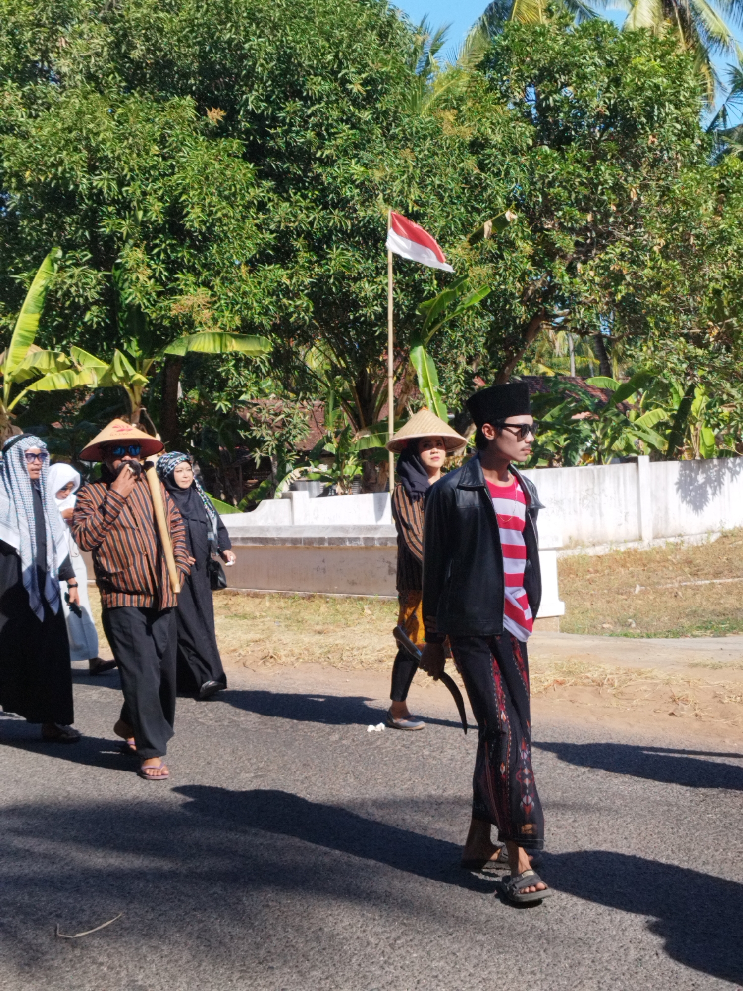 A group of people, some wearing traditional Indonesian attire, walk along a paved road. The group includes individuals in black robes and headscarves, a man in a striped shirt and conical hat, and a young man wearing a black jacket over a red and white striped shirt, black pants, and a peci cap. A red and white flag is visible on a pole in the background, suggesting a possible national or celebratory event. The environment consists of lush green trees and vegetation, with a white wall and buildings in the distance. The lighting suggests it is daytime. The location is identified as Andulang, Indonesia.
