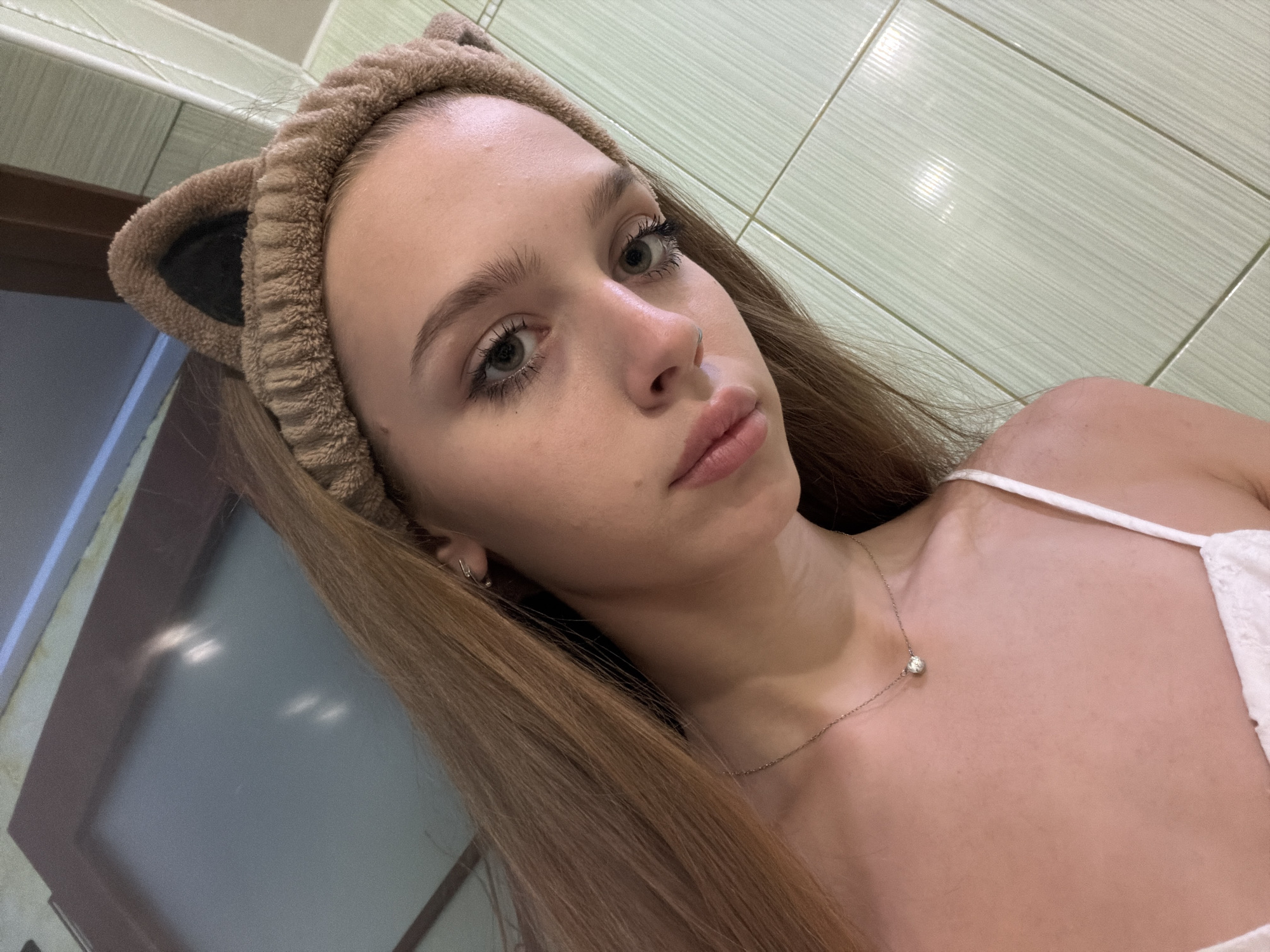 The image is a selfie of a young woman with long, light brown hair, wearing a fuzzy brown headband with cat ears. She has light green eyes, full lips, and is wearing a silver necklace with a small pendant. She appears to be indoors, with light green tiled walls visible in the background. The lighting suggests it could be daytime or evening. Her expression is neutral or slightly serious. The focus is on her face and upper chest.
