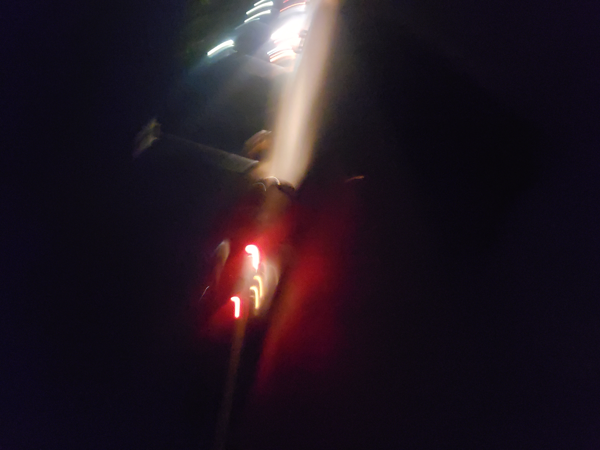 The image is a blurry, nighttime photograph that appears to have been taken from a moving vehicle, possibly in the city of Rido, Nigeria. The scene is dark, with streaks of light indicating motion.
In the foreground, the red taillights of a vehicle are visible, blurred and elongated. To the right, a bright white and yellow light source illuminates a hazy area, suggesting a streetlamp or headlights. There are faint outlines of what could be other vehicles or structures in the background, also blurred. The overall impression is one of a fast-moving scene at night.