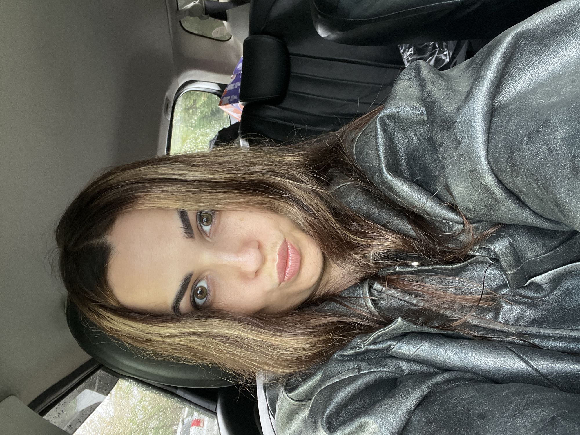 An individual, likely a woman, with long brown hair featuring lighter highlights and brown eyes, is depicted in a close-up, chest-up shot inside a car. Her head is tilted slightly, and she looks directly towards the camera with a neutral to slightly pensive expression, her lips subtly puckered. She is wearing a textured, metallic grey or silver-colored jacket, possibly made of leather or a similar material, with visible folds and wrinkles.
The background shows the interior of a car, including parts of a black car seat or headrest behind her and to her right. Through the window on her left, raindrops are visible, suggesting it is raining or has recently rained. Outside the window, blurry green foliage can be seen, indicating a natural or suburban environment. A small, blurry purple and white object, possibly a package, is also visible through the rear window. The lighting suggests daylight, possibly overcast due to the weather. The individual appears to be taking a selfie or being photographed while in the vehicle. There is no visible text in the image.