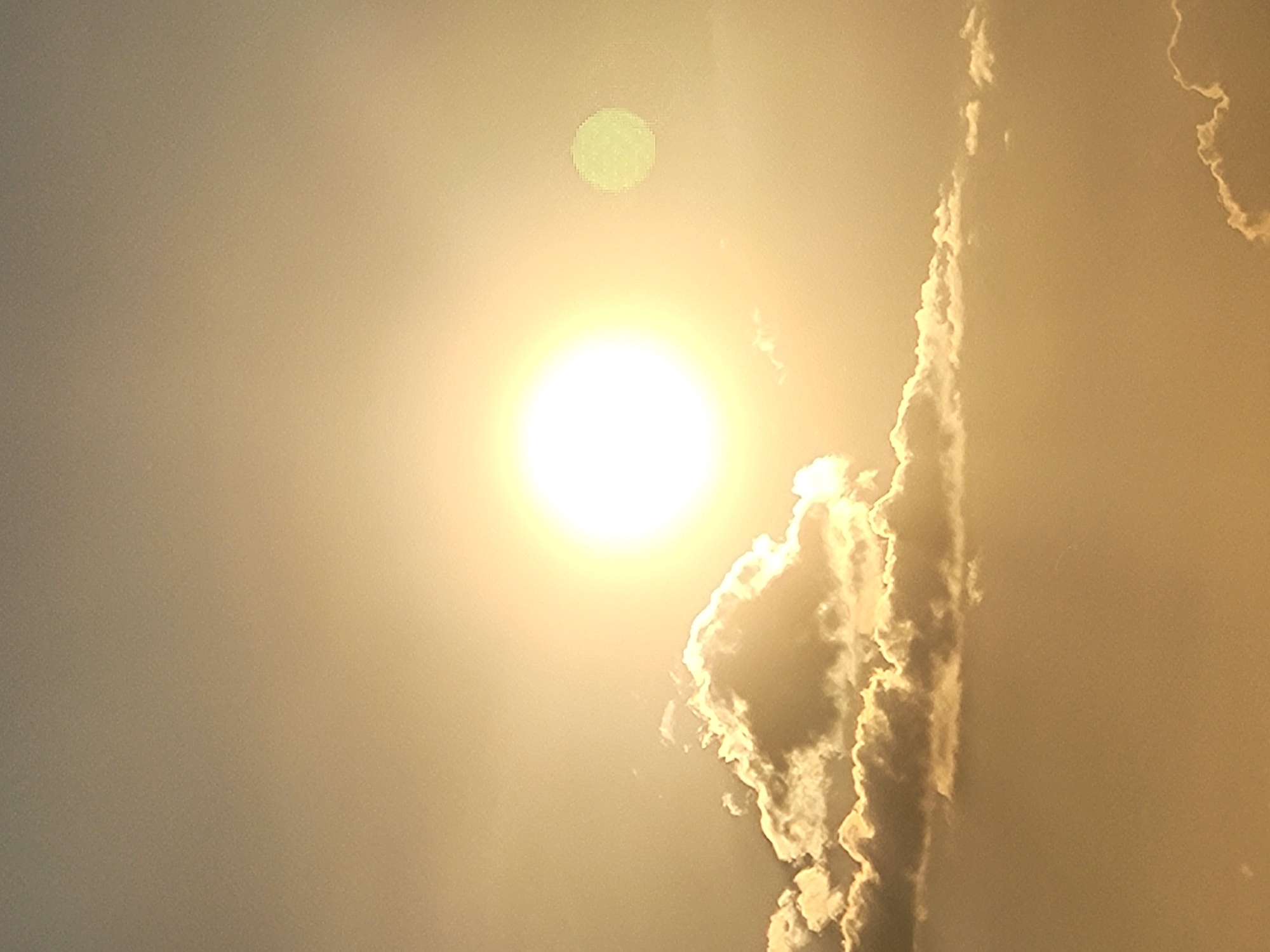 The image shows a bright sun in a hazy, golden sky, with scattered clouds below it. A smaller, less distinct circular object, possibly a lens flare or another celestial body, is visible to the right of the sun. The overall atmosphere is warm and serene. The scene is not identifiable as being in the city of Unknown, Unknown.