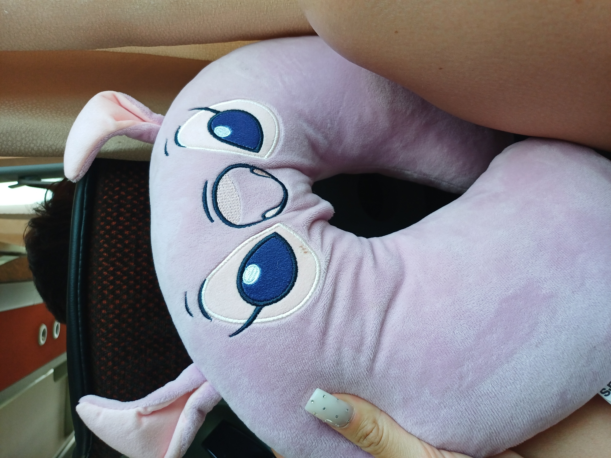 A close-up shot focuses on a pink, plush neck pillow shaped like a character with large blue eyes and pink ears, positioned on a person's lap. The pillow is adorned with embroidered facial features, including eyebrows, eyelashes, and a small mouth. A hand with a nail adorned with rhinestones is visible on the left side of the frame, touching the pillow. The background is blurred, showing hints of a car interior, including seats and a window with curtains. No specific location within Lumina, Romania, is discernible from the image.