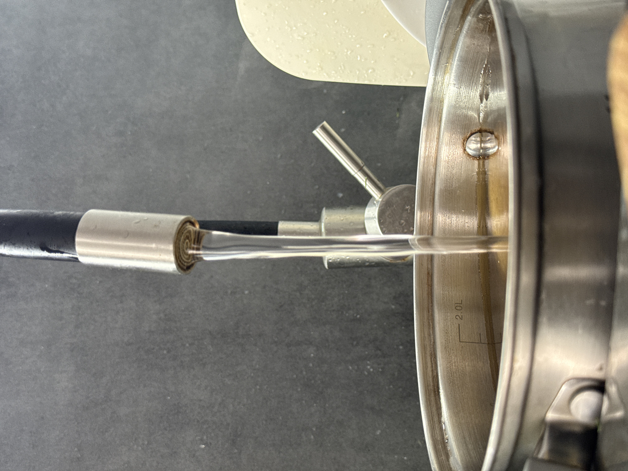 A water hose is filling a stainless steel pot with water on a gray countertop. The pot has a 2.0L fill line visible. The scene is not located in Rostov-on-Don, Russia as there is no evidence in the image to indicate its location.