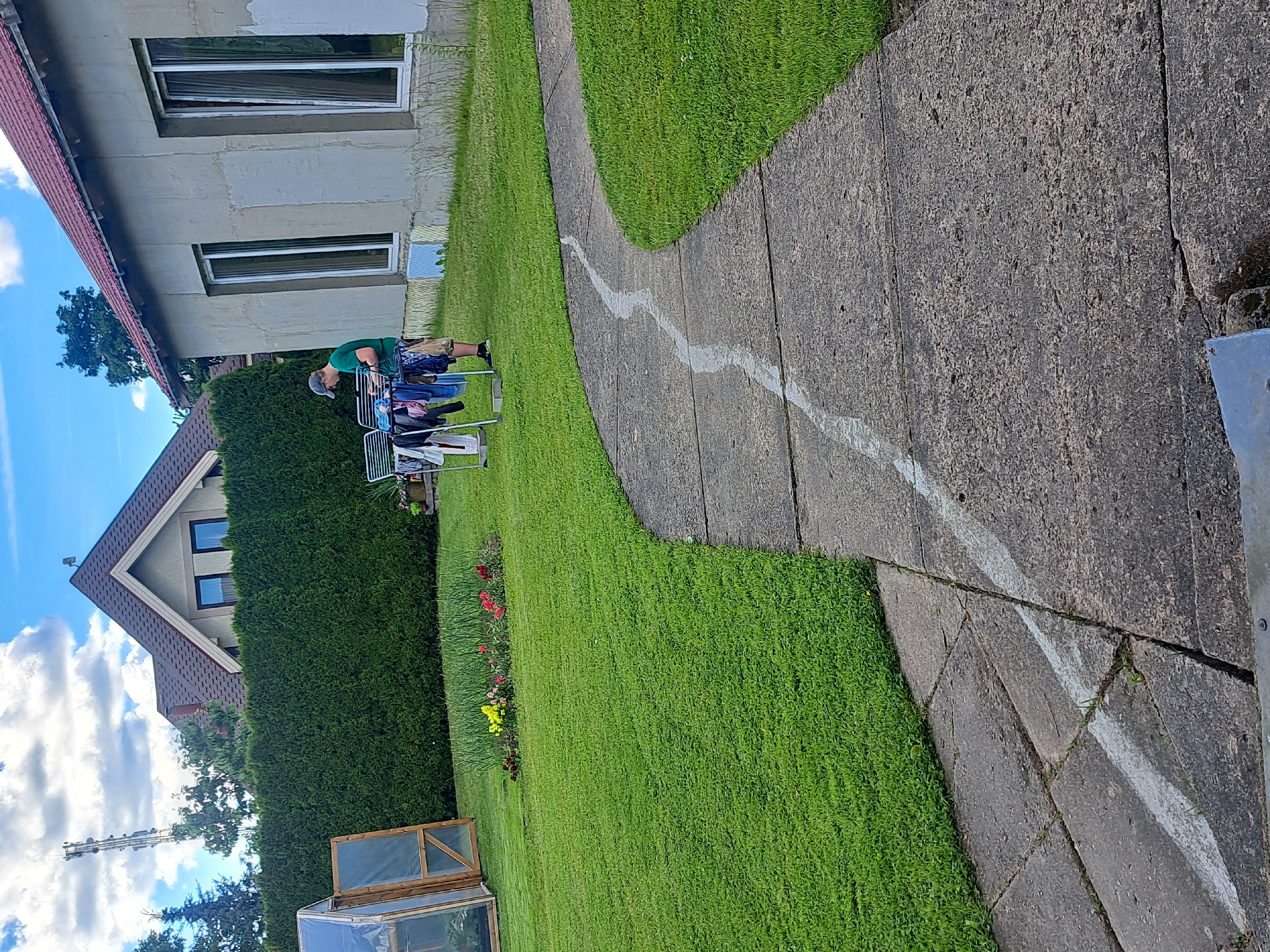 A person wearing a green t-shirt and a blue baseball cap is standing in a yard and hanging clothes on a drying rack. They are facing a building with a light-colored exterior and two large windows. The yard is covered in green grass, and a concrete path with a white stripe runs through it. In the background, there is a tall hedge, a greenhouse, a flower bed, and houses. The sky is blue with some clouds.