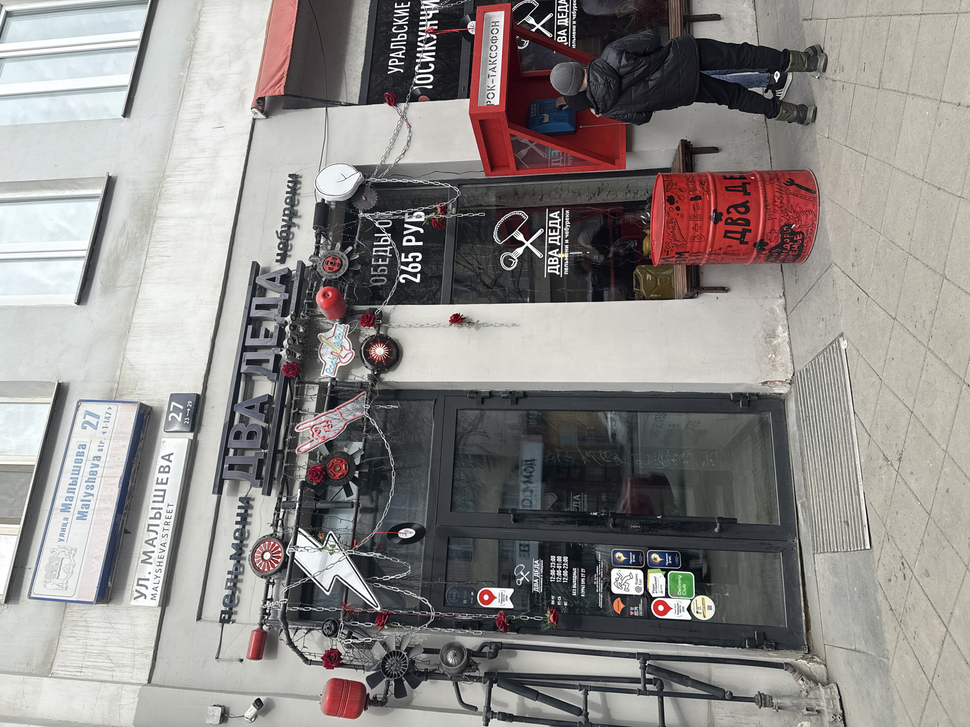 The image shows the facade of a restaurant named "Dva Deda" (Two Grandfathers) in Yekaterinburg, Russia. The restaurant is located at 27 Malysheva Street. The entrance is decorated with industrial-themed items, including gears, pipes, and metal chains, combined with red roses. There is a red barrel with the restaurant's name painted on it, and a red telephone booth labeled "Rok-Taksofon" (Rock-Phonebooth). A person wearing a black jacket and a gray hat is standing near the telephone booth, seemingly interacting with it. Street signs with the name of the street, "Malysheva," are visible on the building. The restaurant's operating hours and delivery service logos are displayed on the glass door.
