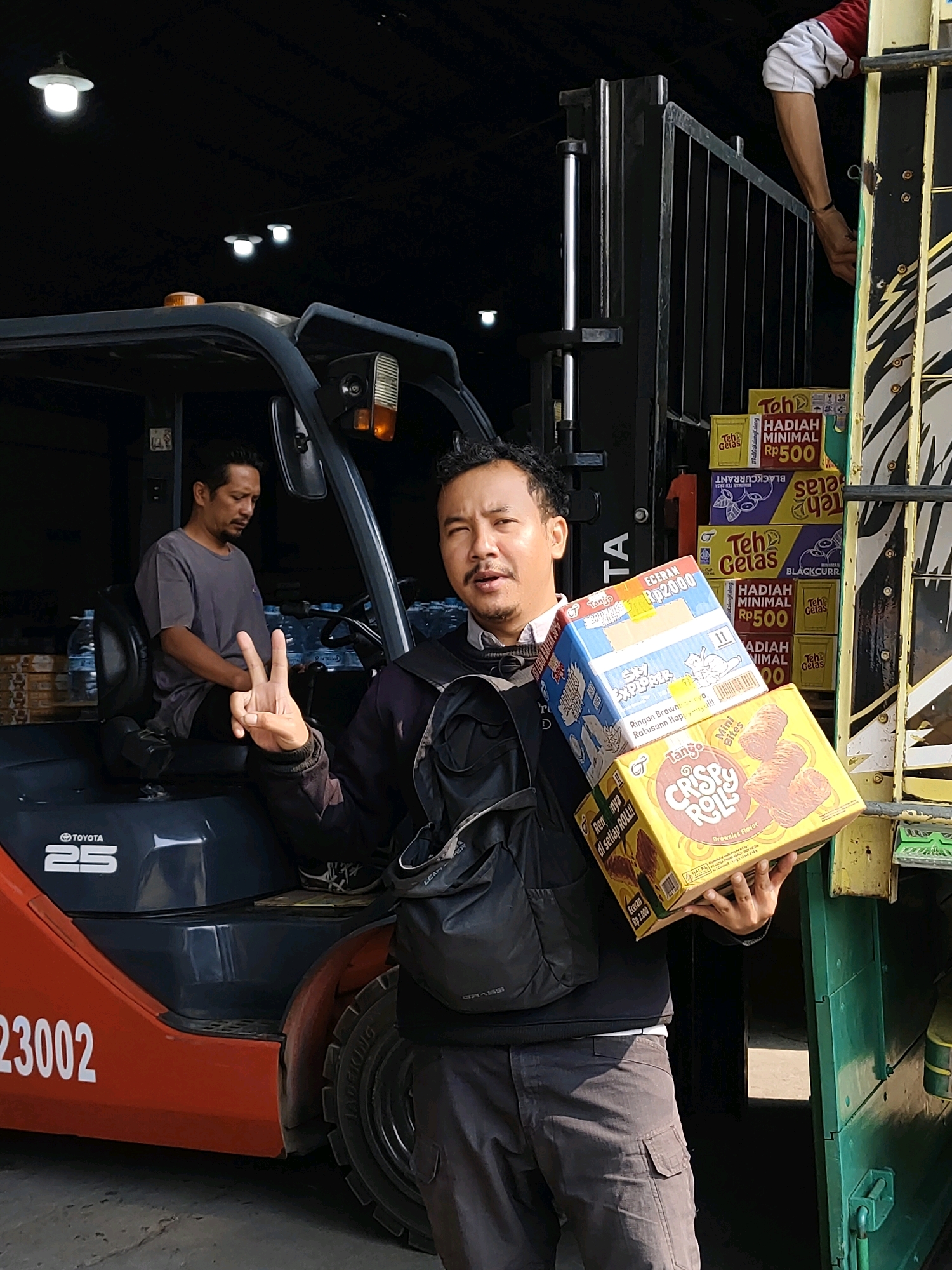 In Probolinggo, Indonesia, a man is holding two boxes of snacks, one labeled "Tango Crispy Roll" and another smaller box with various images and text. He is standing in front of a forklift, which has "TOYOTA 25" and the number "23002" visible on its side. Another man is seated on the forklift, looking down. In the background, stacks of boxes labeled with "Teh Gelas" and "HADIAH MINIMAL Rp500" are visible. The scene appears to be in a warehouse or loading area.