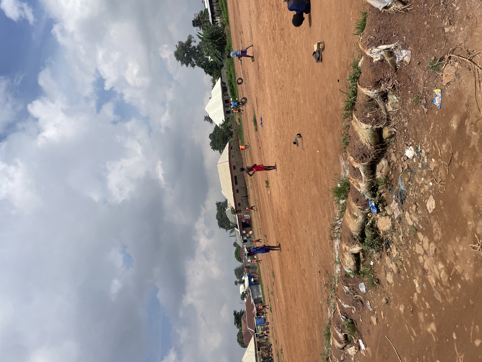 A group of children are playing soccer on a dirt field in Kurunduma, Nigeria. The field is red dirt with some patches of grass and rocks. In the background, there are several houses with corrugated metal roofs, some of which are painted white. The sky is cloudy with some patches of blue. The children are wearing casual clothing and appear to be enjoying themselves. There are also some tires and debris scattered around the field. One child is lying on the ground, possibly resting or injured.