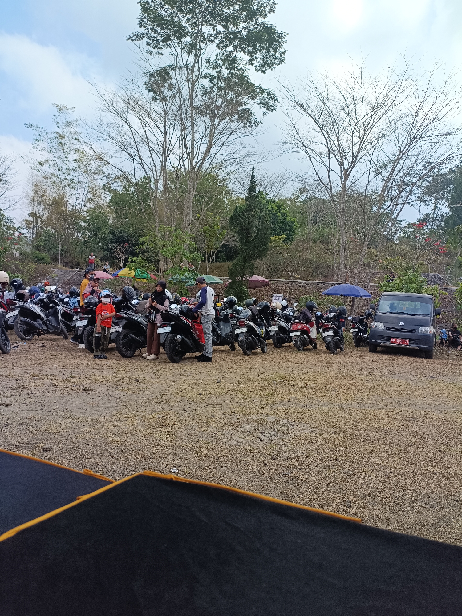The image depicts an outdoor scene in Hargobinangun, Indonesia, with a large number of motorcycles parked in an open, dusty lot. Several individuals are present, some standing near the motorcycles, others in the background, and a few sitting on the ground. A dark-colored van is parked on the right side of the lot. In the foreground, there is a dark fabric with an orange trim, possibly a cover or a piece of equipment. The background features trees, vegetation, and a slightly overcast sky. The overall impression is of a gathering or event where many people have arrived by motorcycle.