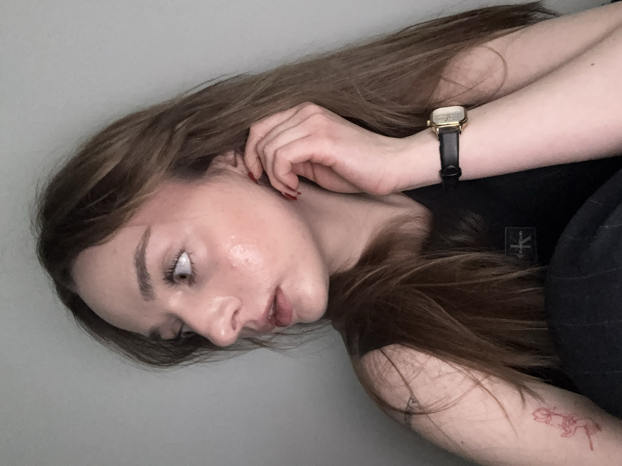 A young adult female lies on a gray surface, presumably a floor or bed, angled slightly towards the camera. She has long brown hair and light skin. Her head is turned to the left. Her right hand is near her right ear, and she is wearing a wristwatch with a black band. Her left arm is bent across her body, and a faint red tattoo is visible on her left forearm. She is wearing a black top. The location is unknown.