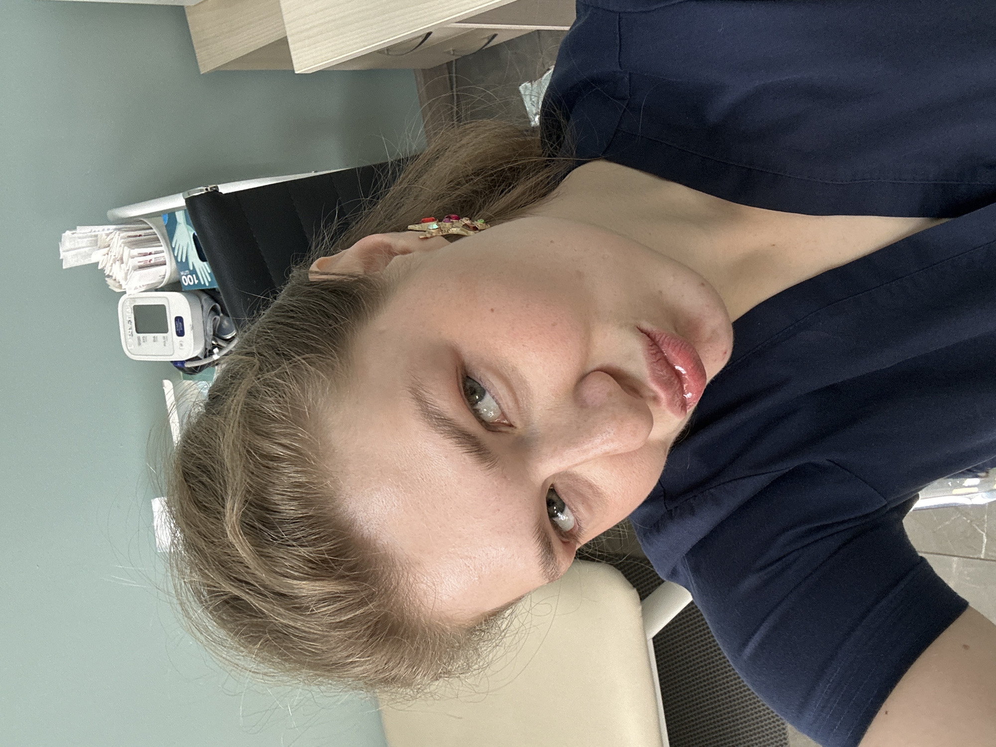 A close-up, slightly angled photograph shows a fair-skinned woman with blonde hair, wearing a dark blue garment, possibly medical scrubs. She has light eyes and her lips are glossy. She is indoors, seemingly in a medical setting, identifiable by the medical equipment visible in the background, including a blood pressure monitor and disposable gloves. The location is unknown. There is no indication this image was taken in Kazan, Russia.