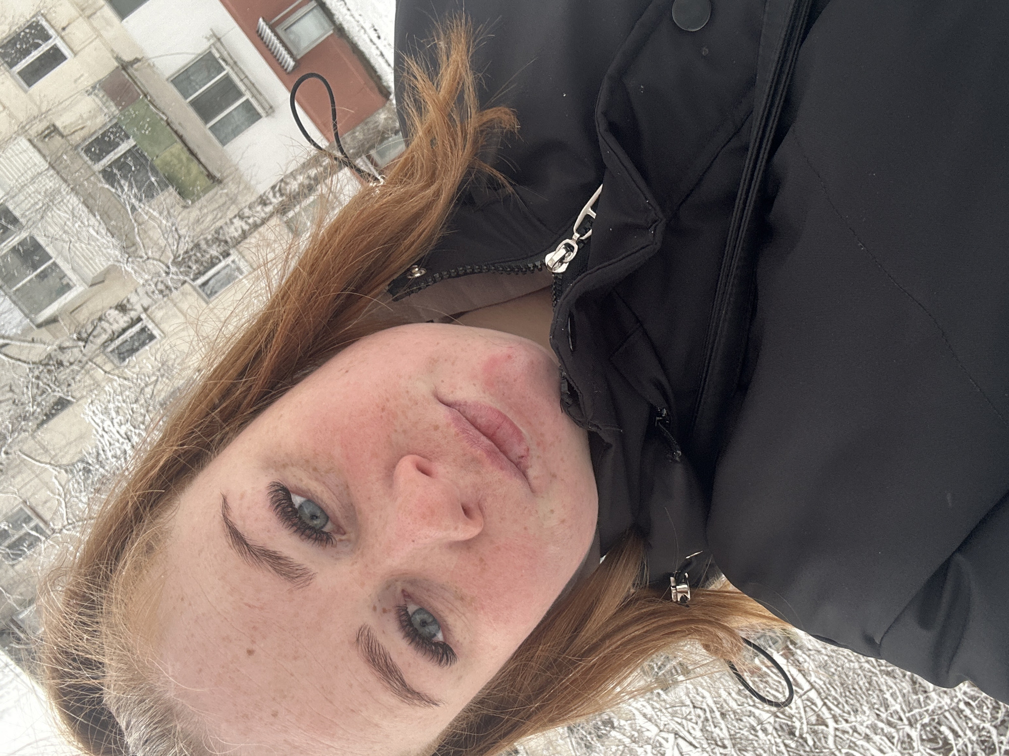 A fair-skinned, red-haired woman with freckles and blue eyes is the focal point of this outdoor photograph. She is wearing a black winter coat with a zipper and buttons. The background features a multi-story building with multiple windows, potentially an apartment complex. The presence of snow on the ground and bare trees suggests a winter season. Given the context, the location is likely within the city of Chaikovsky, Russia.