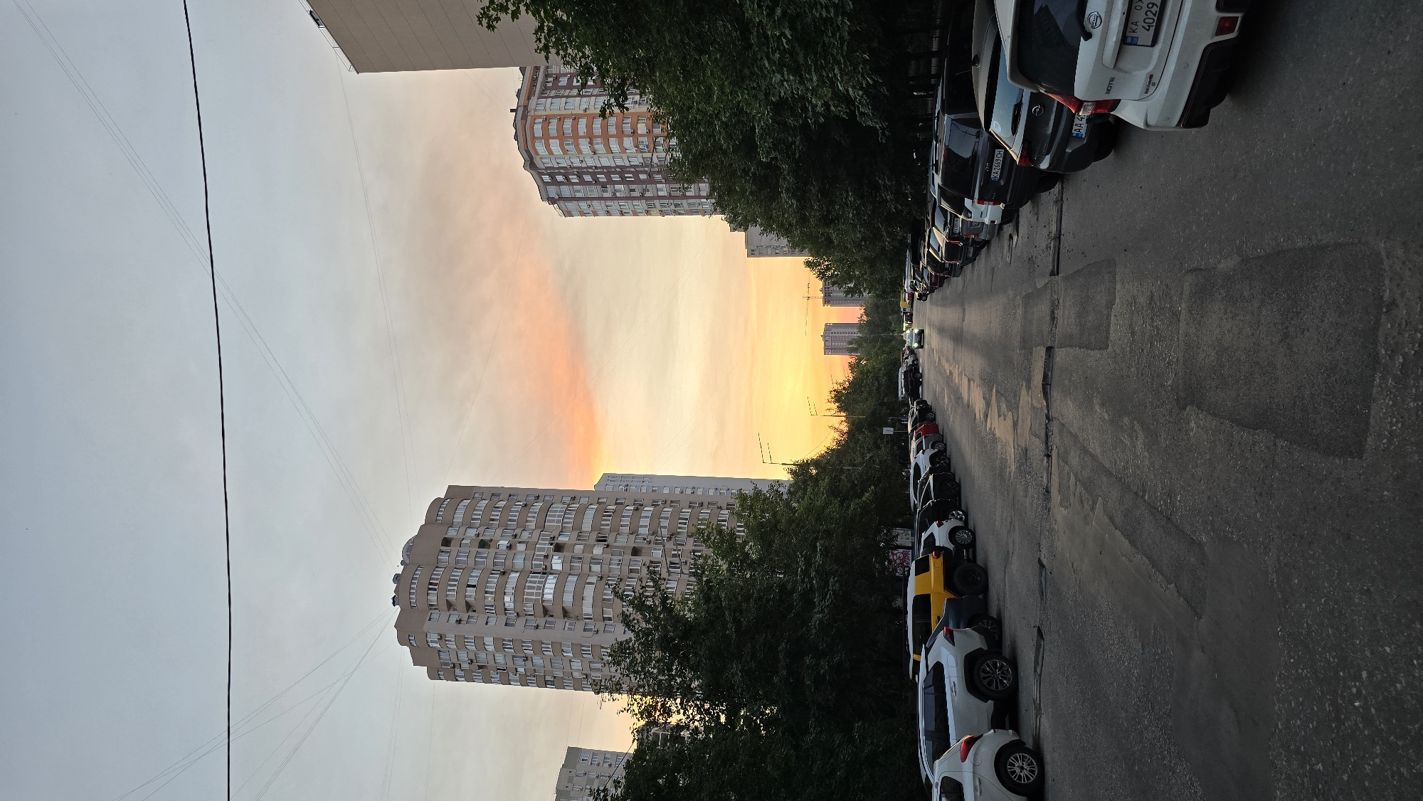 The image depicts a street scene in Kyiv, Ukraine, at dusk. Two tall, multi-story apartment buildings dominate the left and right sides of the frame. The sky above displays a gradient of colors, transitioning from a pale blue at the top to warm orange and yellow hues near the horizon, indicating the sunset.
A paved road runs through the center of the image, lined on both sides with parked cars. The road surface appears to be asphalt and shows signs of wear, with several patched areas. Trees with dense green foliage are situated between the road and the apartment buildings.
In the distance, more apartment buildings can be seen, appearing smaller due to their distance. A single vehicle is visible driving down the road, with its headlights on. The overall impression is that of a residential urban environment during the evening.