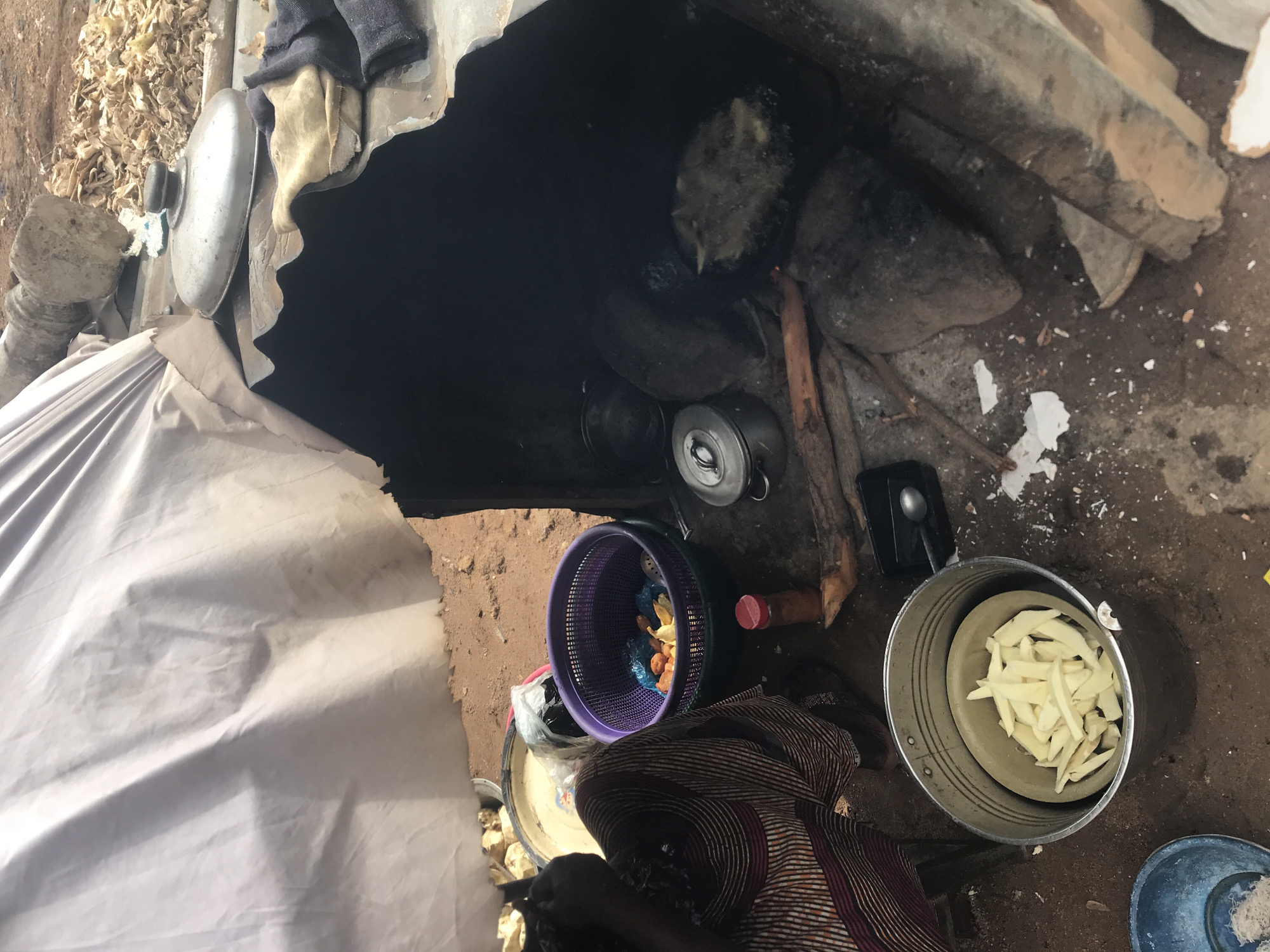 An overhead shot shows a cooking scene in an outdoor, rustic environment, likely in Rafin Par, Nigeria. A woman wearing a vibrantly patterned dress is seated to the left, with her back partially to the viewer. She is next to a large, grey metal bucket containing a lighter-colored bowl filled with sliced, pale yellow food items, possibly plantains or yams. In front of the bucket, a small blue bowl is partially visible. To the right of the woman is a purple basket filled with food items, including some orange and yellow pieces. A small red-capped bottle sits on the ground beside the basket.
The central focus of the image is a makeshift outdoor oven constructed from corrugated metal. This oven is open on one side, revealing a dark interior where a pot is actively cooking over a fire. The pot contains bubbling oil with food items being fried. Several metal pots and lids are scattered around the oven. The ground is dirt, with some scattered debris including wood, white paper fragments, and possibly dried food matter. In the background, a large, light-colored tarp is draped over a structure, and a pile of dried leaves or husks is visible. A large, cylindrical concrete object is also present in the background. The overall impression is one of a resourceful and active cooking environment.