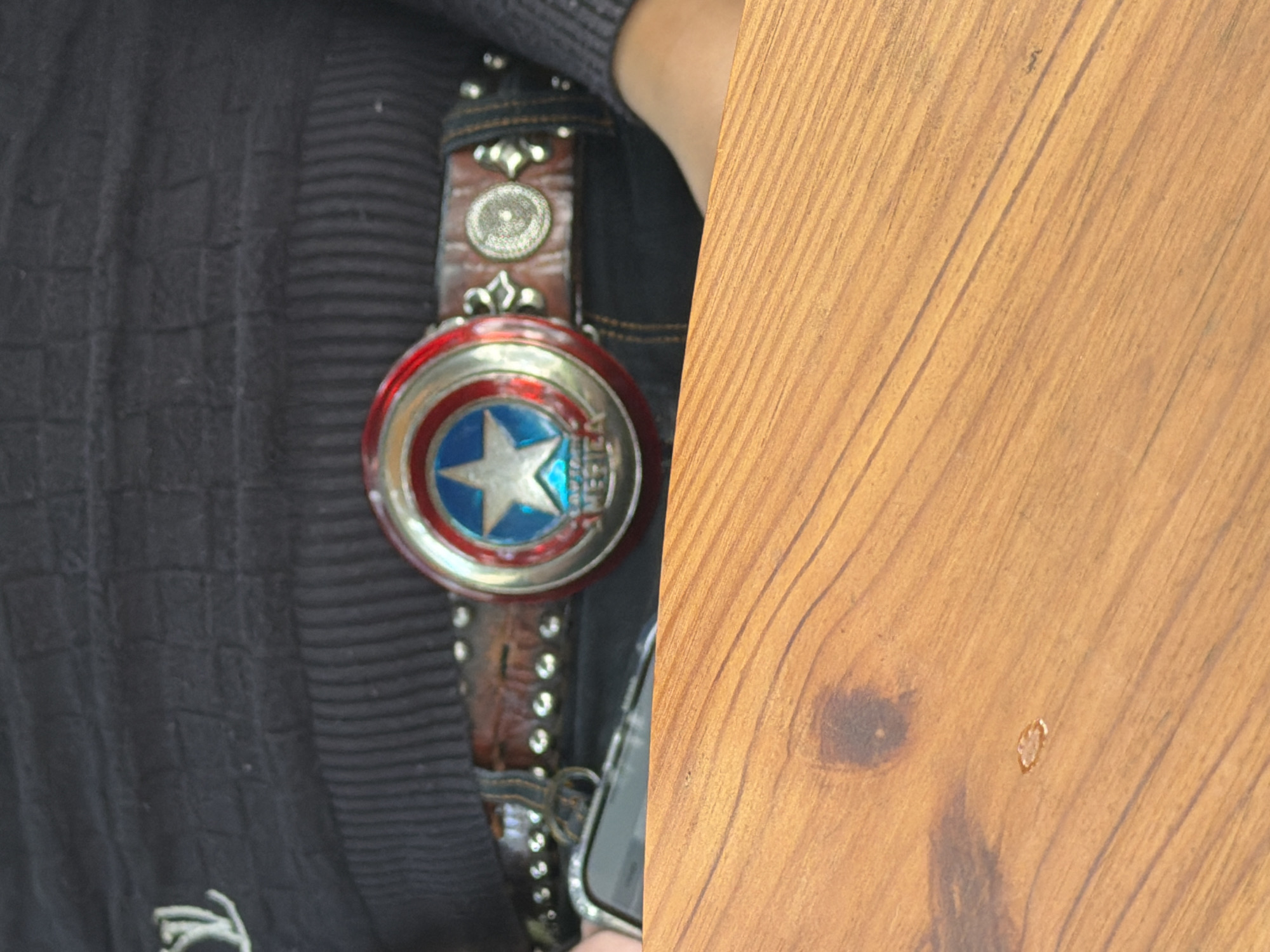A person is wearing a black sweater over a pair of black pants and a belt with a Captain America shield buckle. The belt is made of brown leather and adorned with silver studs. The person is holding a smartphone. The background is a wooden surface. The image is taken at ground level.