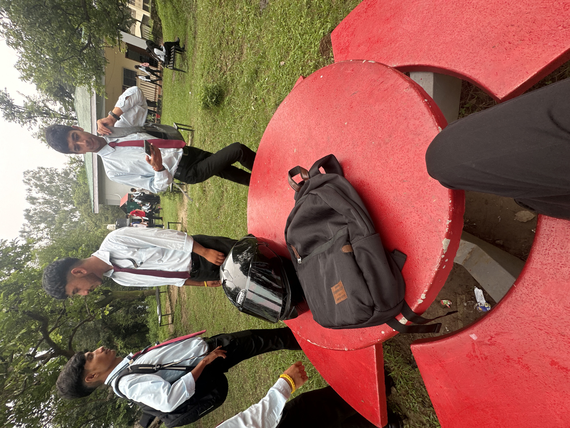 In an outdoor setting, two young men in light blue collared shirts and burgundy ties stand near a red picnic table. One man, facing left, wears a backpack and has his left hand on his hip. The other man, facing right, holds a cell phone and adjusts his backpack strap over his shoulder. On the picnic table are a black helmet and a black backpack. Another young man, wearing a light blue collared shirt and burgundy tie, stands with his back to the camera to the left of the table, wearing a backpack. In the background, a building with a green roof is visible, and several other individuals are scattered around the grassy area. The environment appears to be a park or campus with trees and overcast skies.