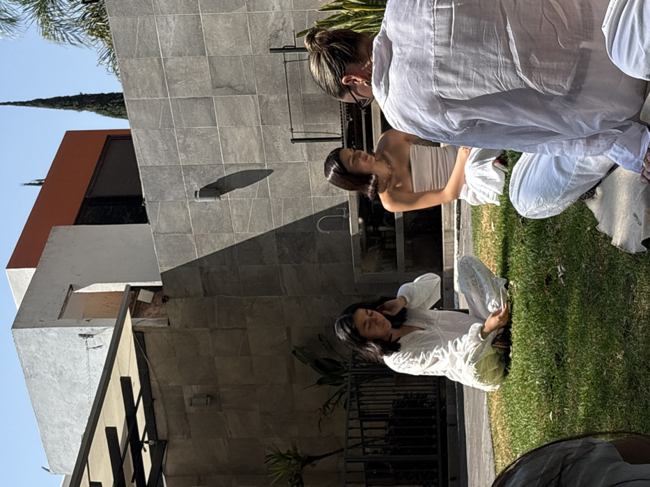 The image displays four female individuals seated on a sunny grass lawn within a modern residential courtyard or backyard, presumably located in Puebla, Mexico.
The foreground shows the left shoulder and arm of an individual clad in a light-colored sleeve. Adjacent, a young female with dark, shoulder-length hair wears a white embroidered long-sleeved top and matching patterned pants. Her right hand is raised towards her ear, while her left hand holds a small white object, and her gaze is directed downward. Centered in the midground, another young female with dark, short, styled hair wears a strapless light-colored top and white pants; she is gazing towards the right. To her right, an adult female with light-colored hair styled in a bun, wearing glasses and a loose white linen-like shirt and pants, sits on a light-colored mat with her back partially towards the camera. All individuals are seated cross-legged.
The background features a multi-level building constructed with grey tiled walls, white stucco sections, and an upper-level orange facade element. Dark-tinted windows are visible. Against the grey tiled wall, an outdoor kitchen area includes a sink faucet and a metal barbecue grill structure. Two wall-mounted light fixtures are visible on this wall. An overhead awning structure with a black metal frame and light-colored fabric cover is positioned on the left side of the scene. Beyond the building walls, tall, slender conifer trees are visible against a clear blue sky. Bright sunlight casts distinct, sharp shadows from architectural elements across the tiled wall and grass.
