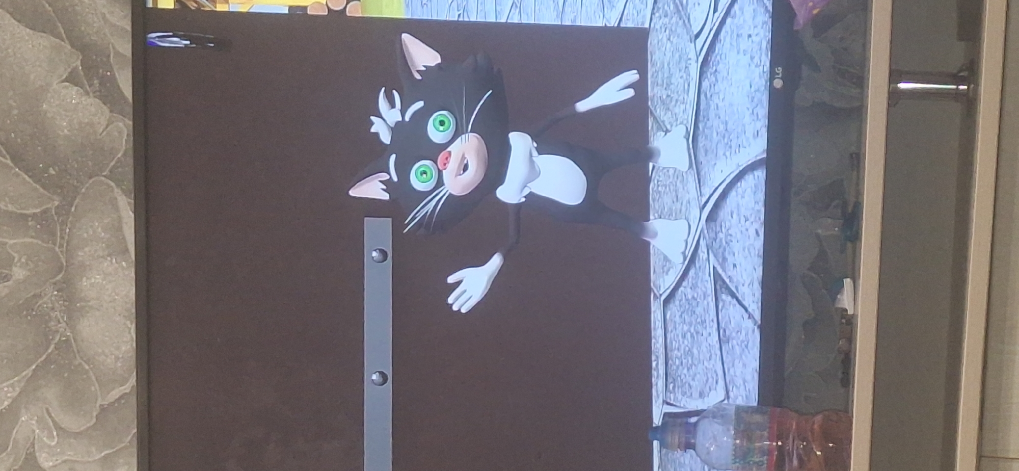 This image shows a cartoon black and white cat with green eyes and pink nose, waving its left hand. The cat is standing on a patterned gray surface. Behind the cat, there is a dark brown wall with a horizontal metal bar featuring two circular indentations. In the lower left corner of the image, a clear plastic bottle filled with a yellowish-orange liquid is visible. The background above the wall appears to be a textured wallpaper with a floral pattern. The scene is captured on a screen, as indicated by the LG logo in the bottom right corner of the screen. The location is not identifiable as Bila Tserkva, Ukraine based on the provided visual information.